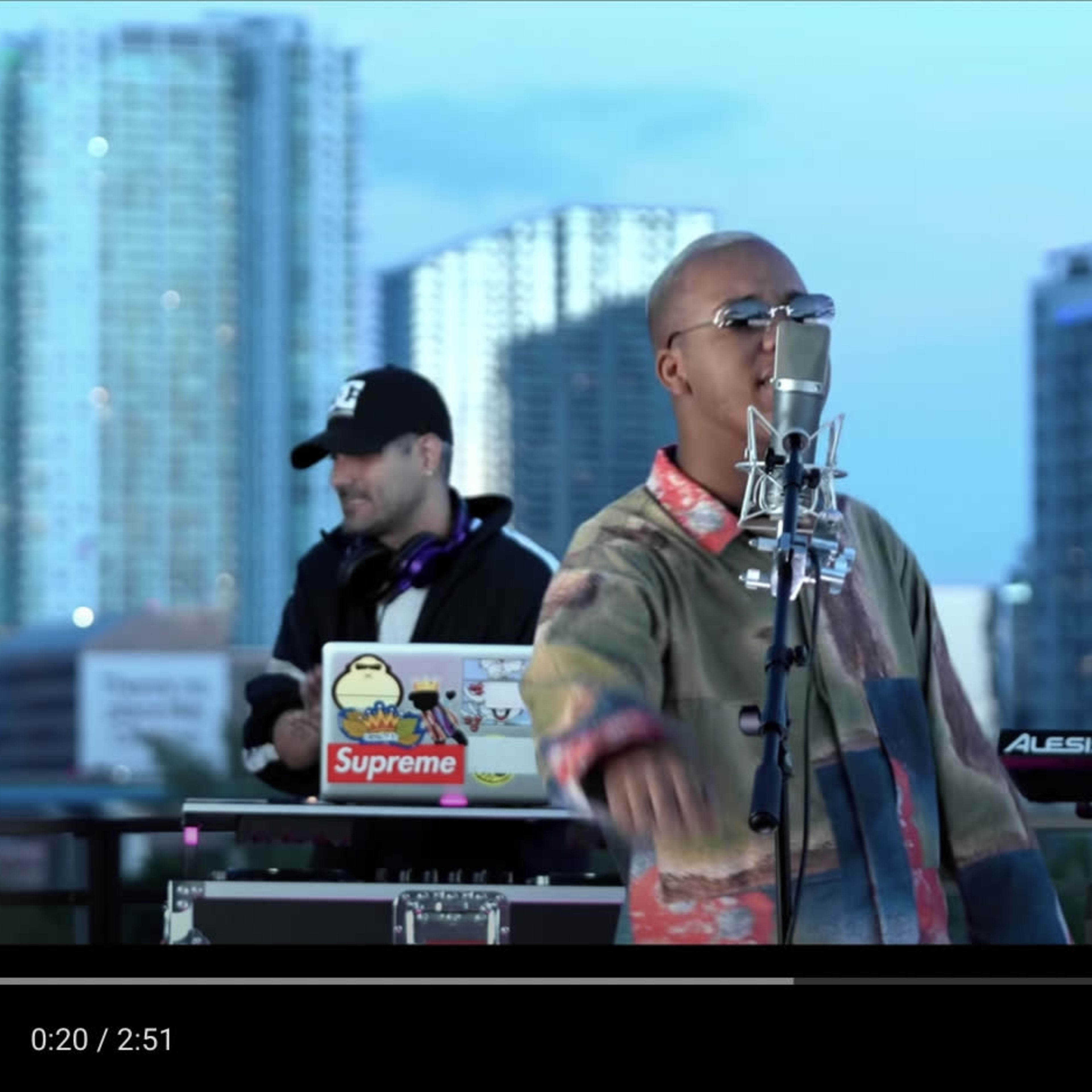 A vocalist sings into a microphone while two musicians play instruments in an urban setting with skyscrapers in the background.
