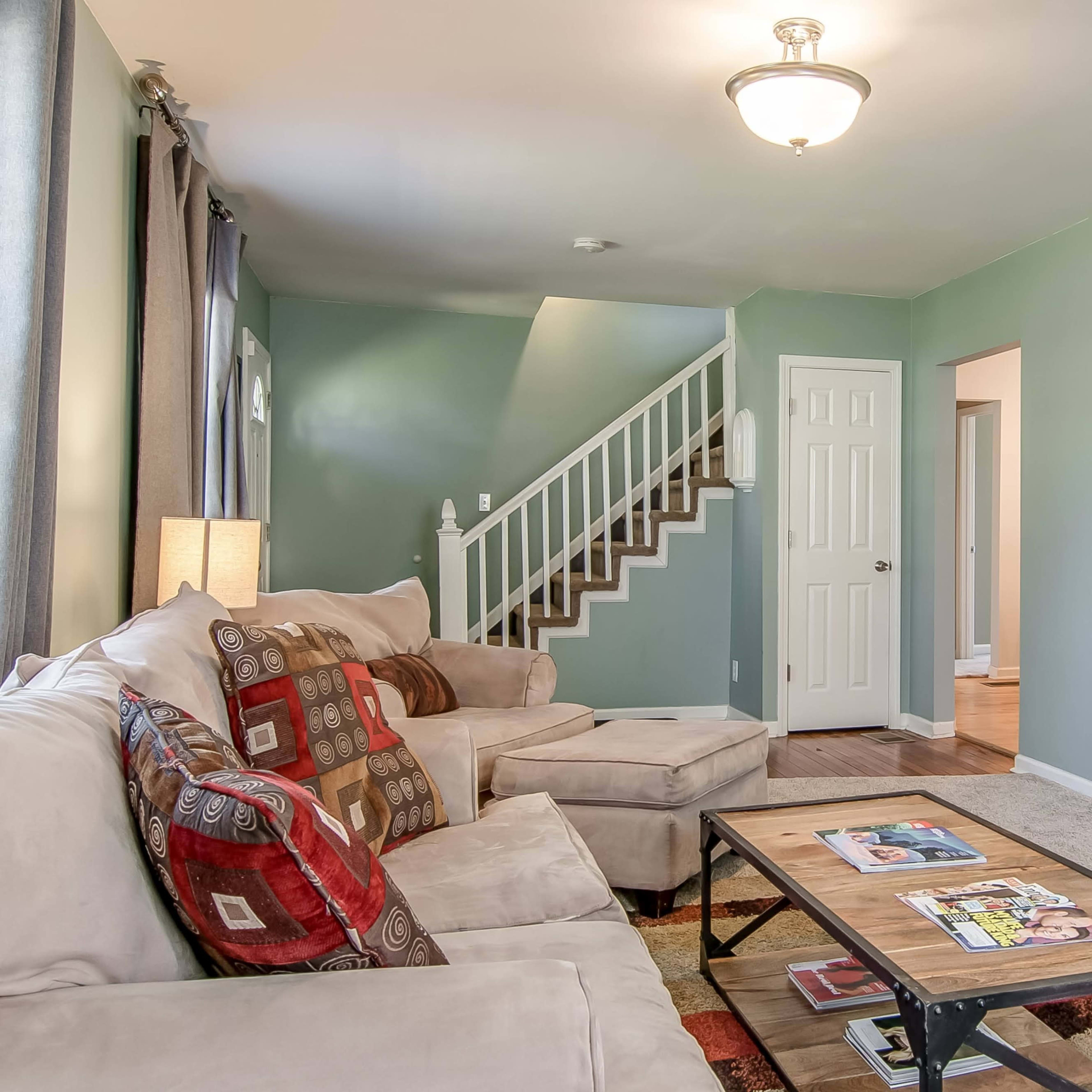A cozy living room features a beige couch, a wooden coffee table with magazines, and a TV stand against a light green wall, with a staircase leading to an upper level.
