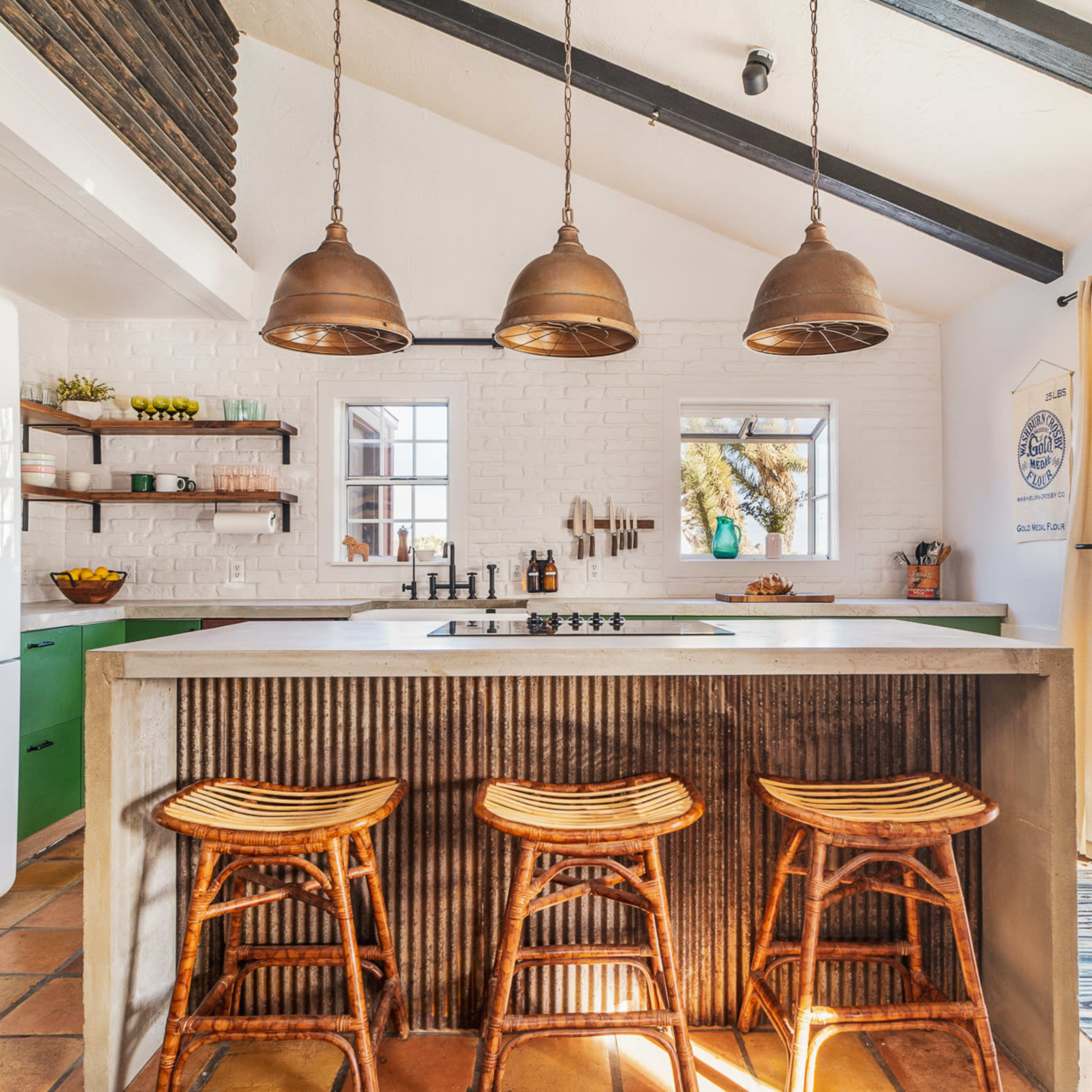A modern kitchen features a concrete island with three wooden stools, pendant lights, and large windows overlooking an outdoor area.
