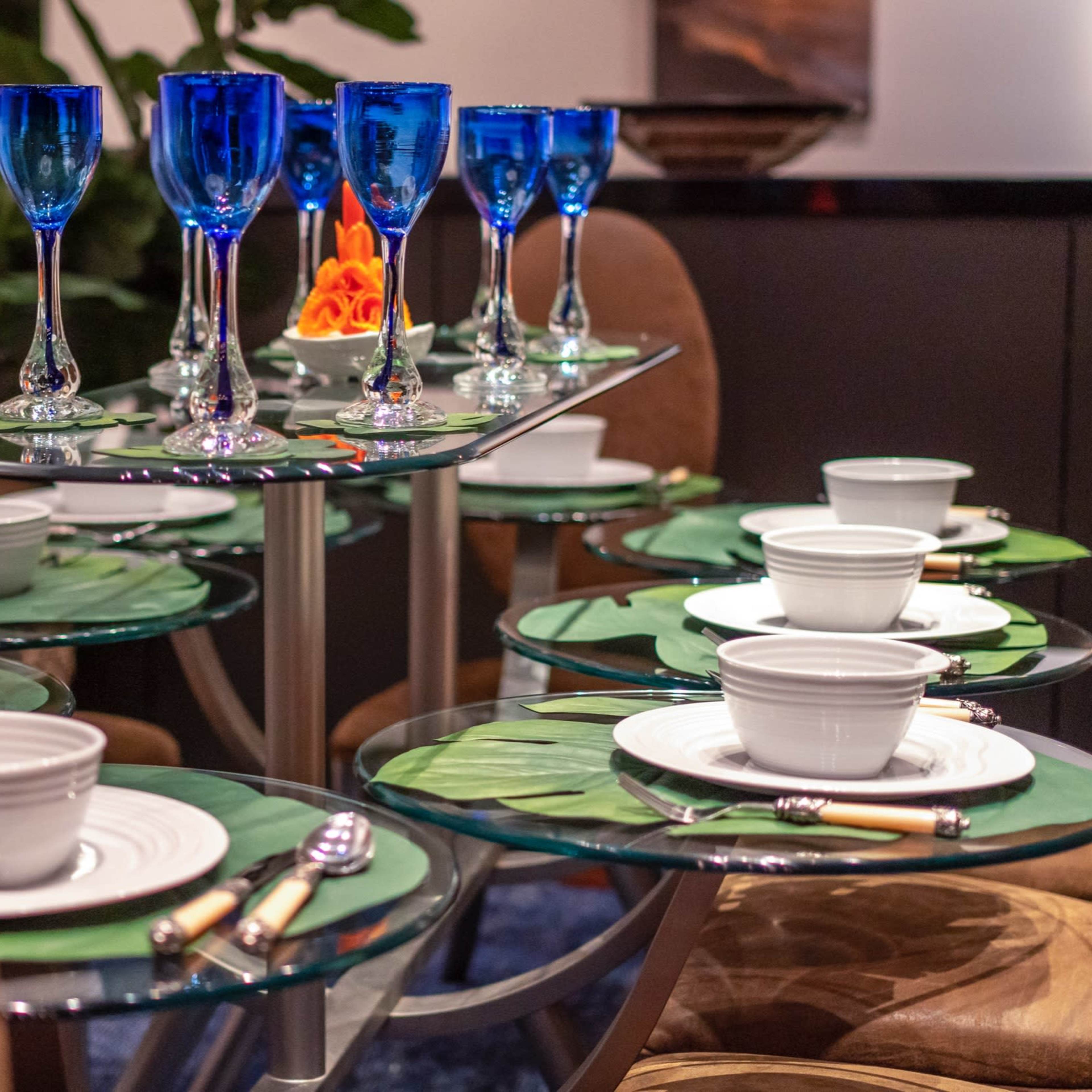 A dining table is set with clear glass plates, white bowls, and blue goblets, accompanied by decorative green leaves and silver utensils.