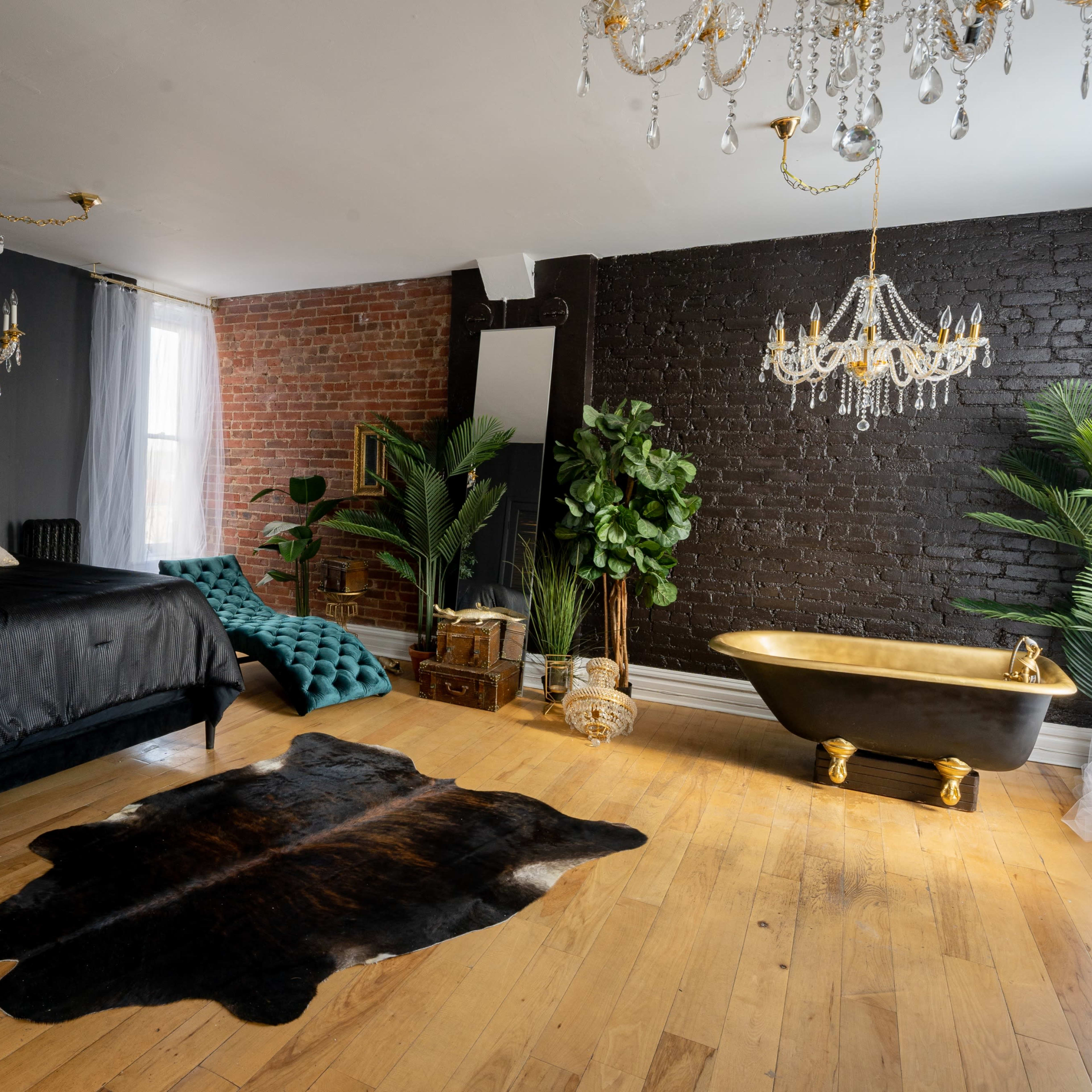 The room features a black velvet bed, a gold-accented freestanding bathtub, and a combination of exposed brick and dark walls, with greenery and decorative chandeliers enhancing the space.