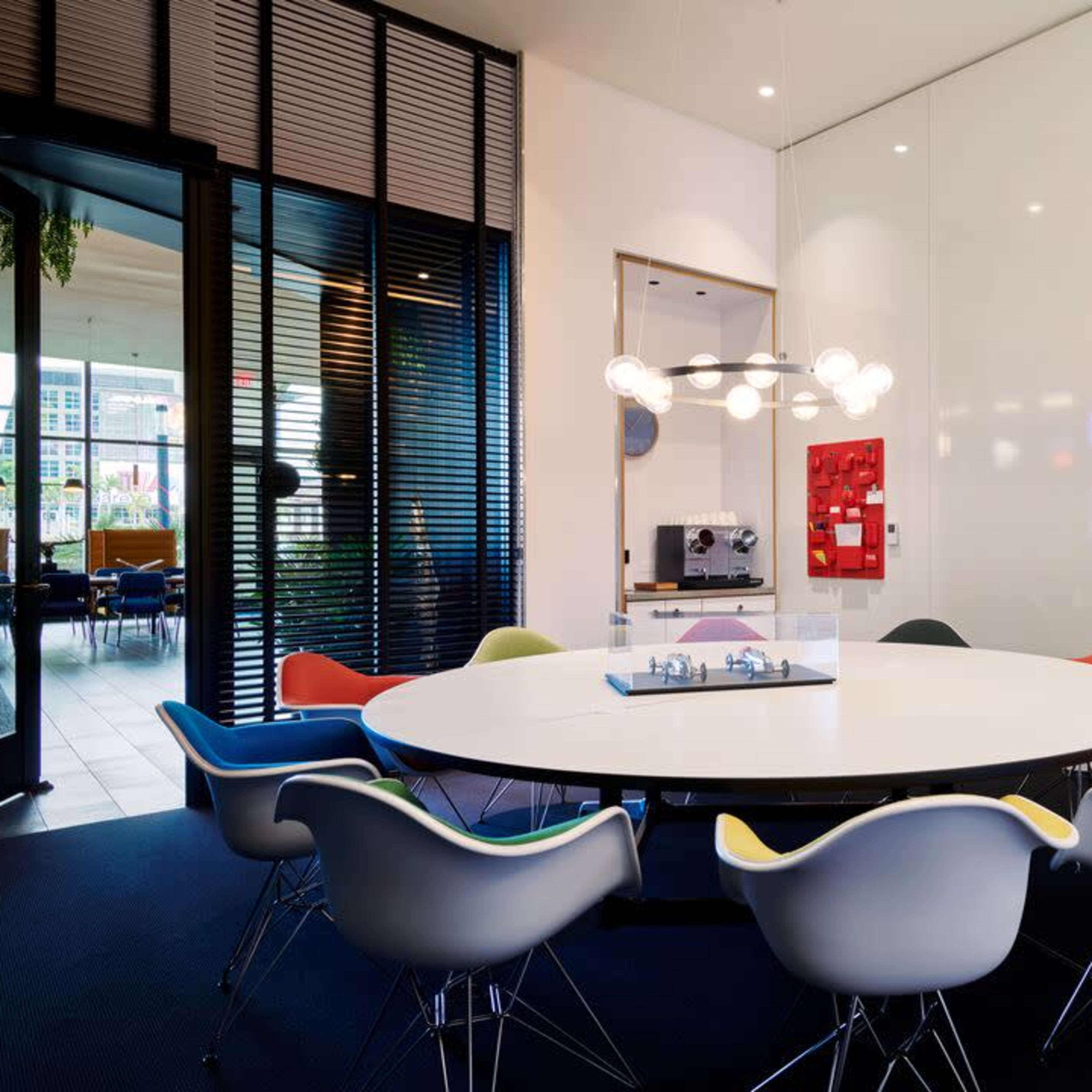 A circular conference table surrounded by colorful chairs is set in a modern meeting room with large windows and minimalistic decor.