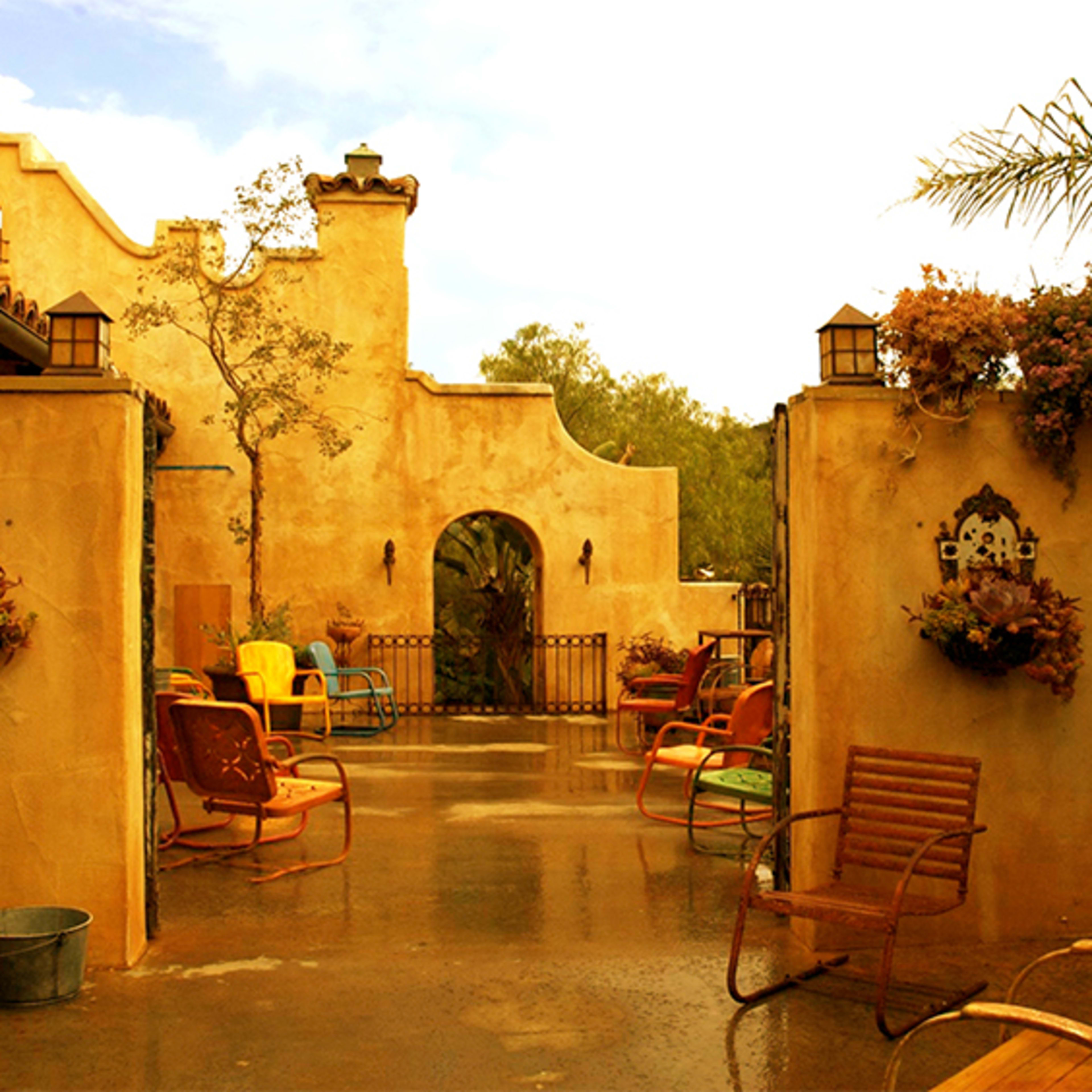 The scene features a courtyard with colorful chairs and potted plants set against a textured, earth-toned wall and an arched entrance.
