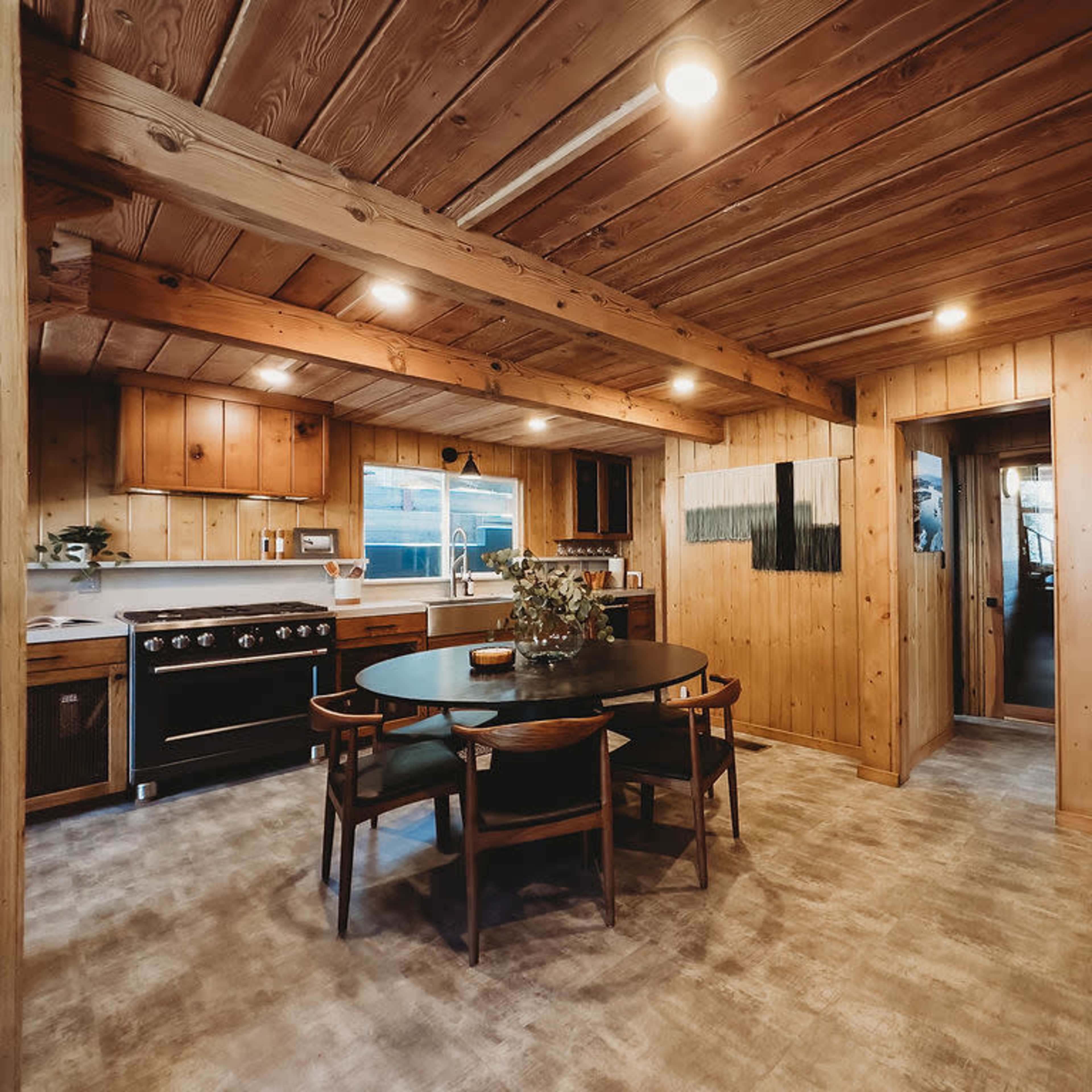 A wooden-finished kitchen and dining area featuring a round dining table, a large stove, and a doorway leading to another room.
