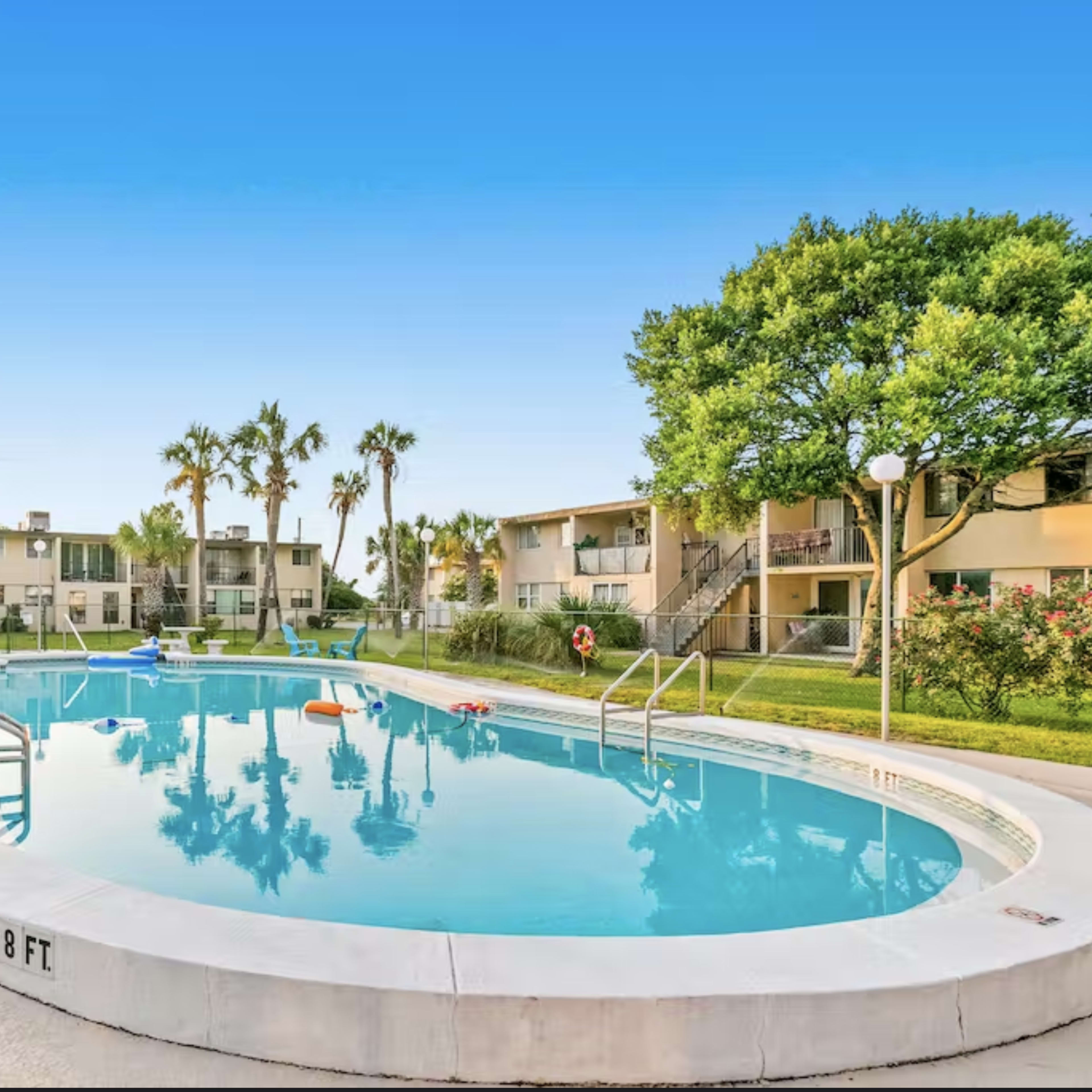 A swimming pool surrounded by palm trees and low-rise apartment buildings under a clear blue sky.