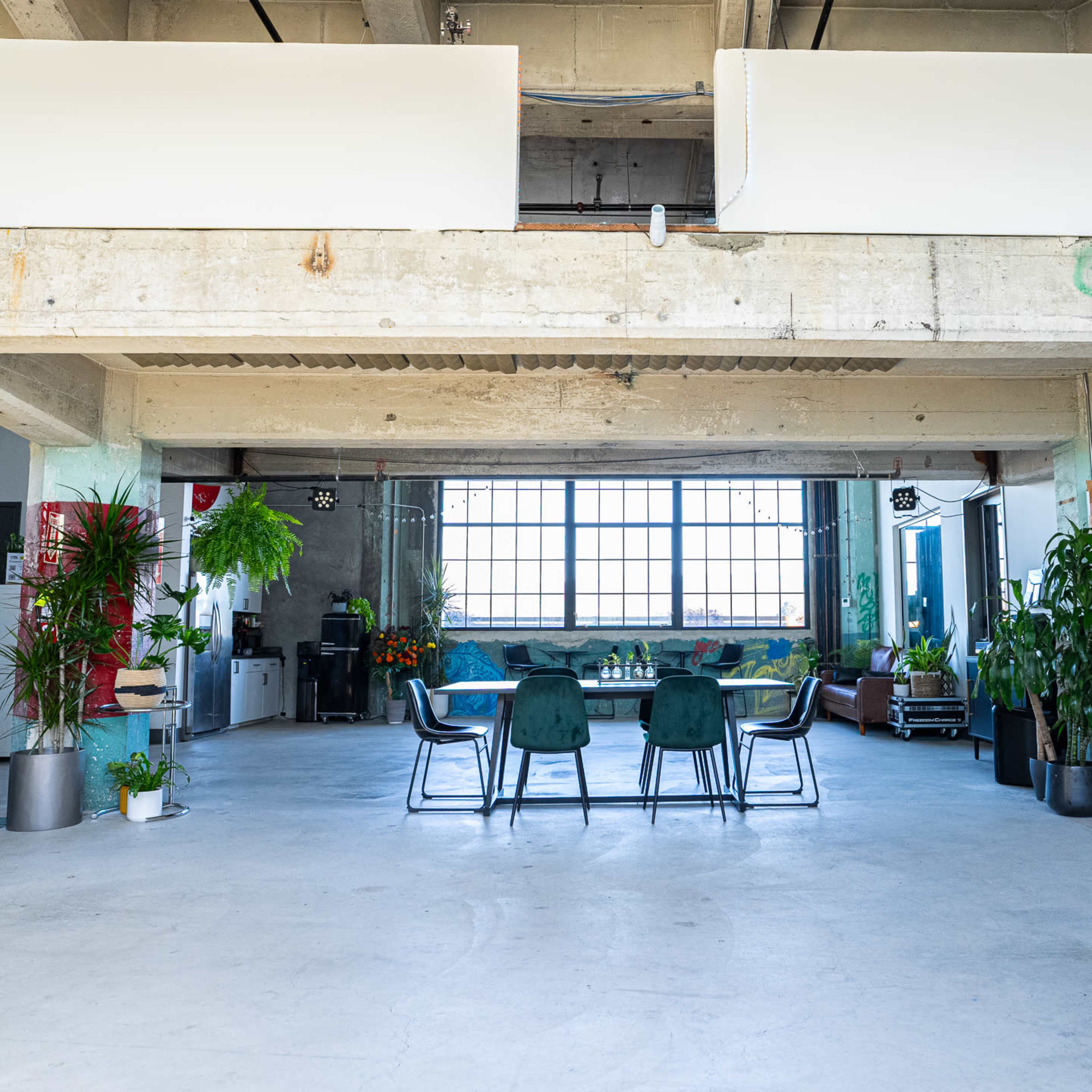A spacious modern loft featuring a large open area with a dining table and chairs, alongside various plants, and large windows that provide natural light.
