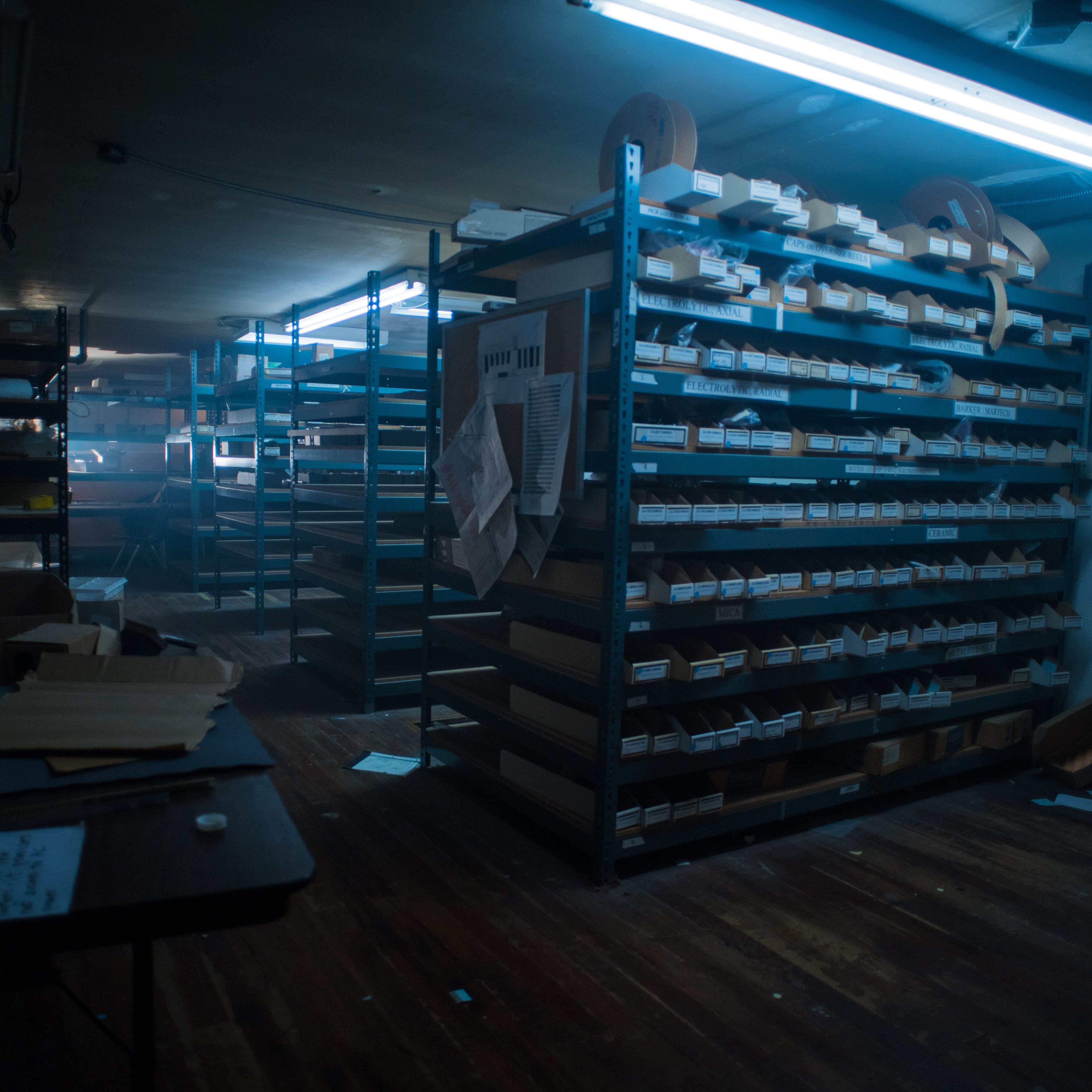 A dimly lit storage room filled with metal shelving units stacked with various labeled boxes and scattered papers on a wooden floor.