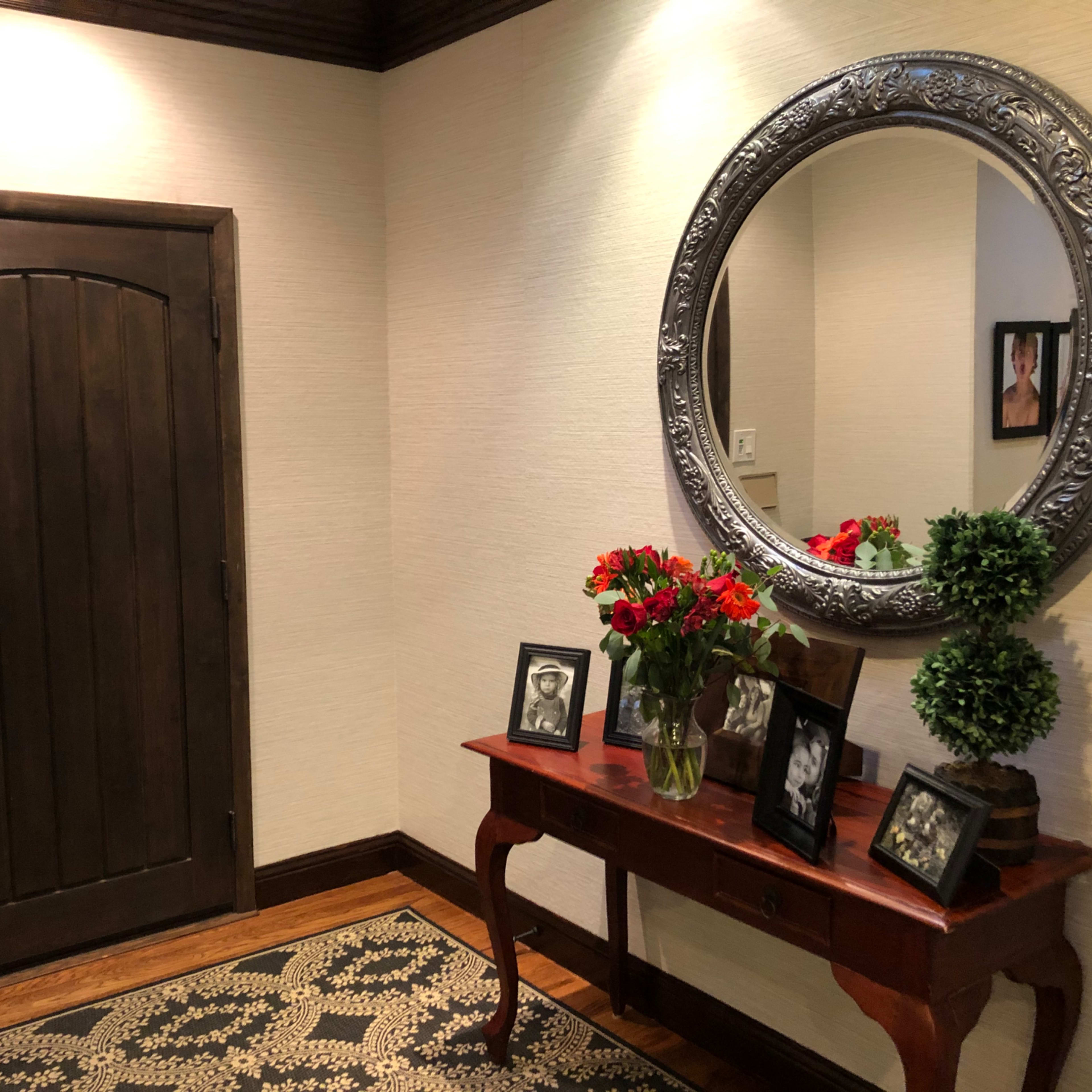 A hallway entrance featuring a round silver mirror above a wooden console table adorned with framed photos and a vase of flowers, with a dark wooden door to the left.