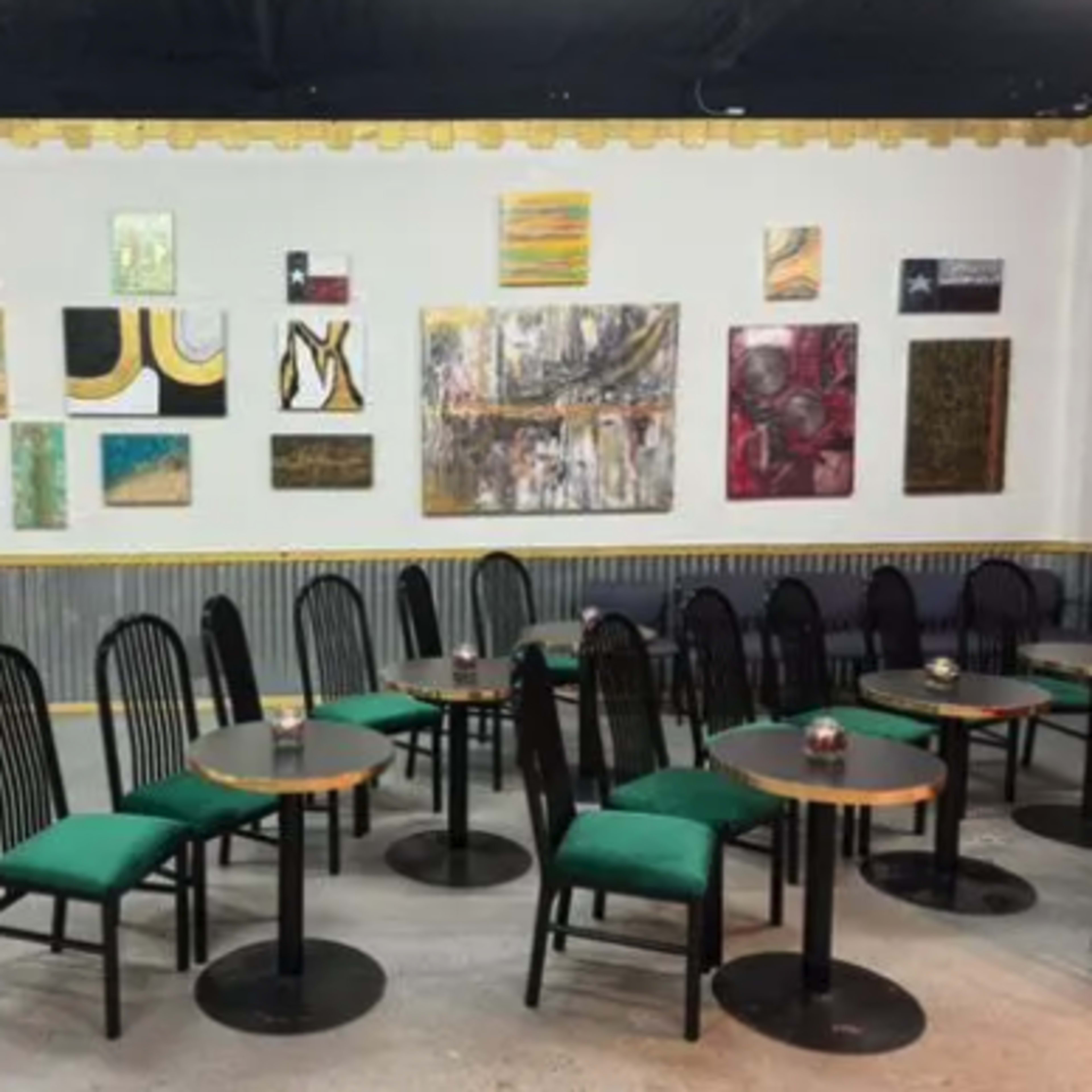 A cafe interior featuring several small black tables with green chairs, set against a wall adorned with various framed paintings.
