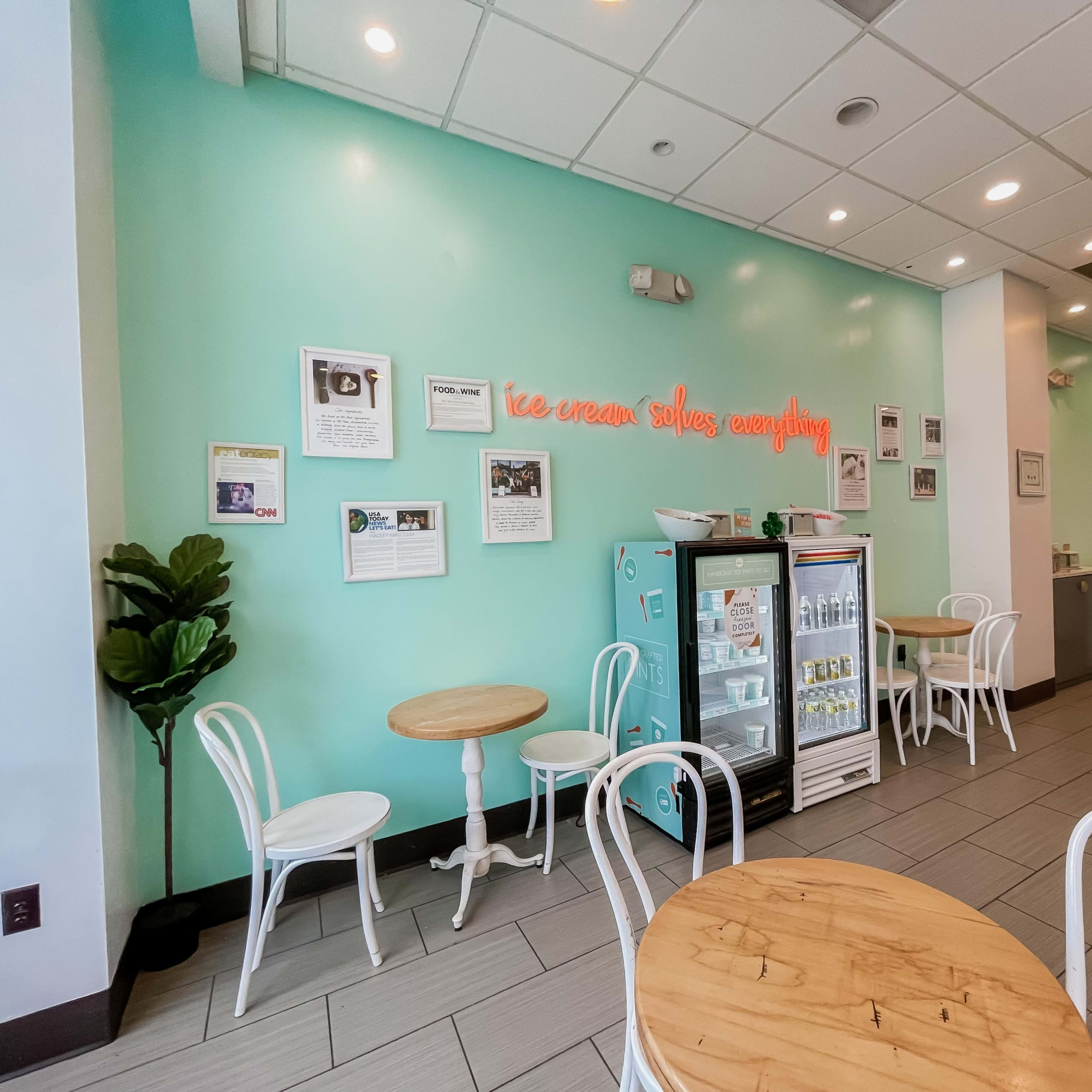 The scene shows a brightly colored ice cream shop with light teal walls, wooden tables, and white chairs, featuring a display case and various framed photos on the walls.