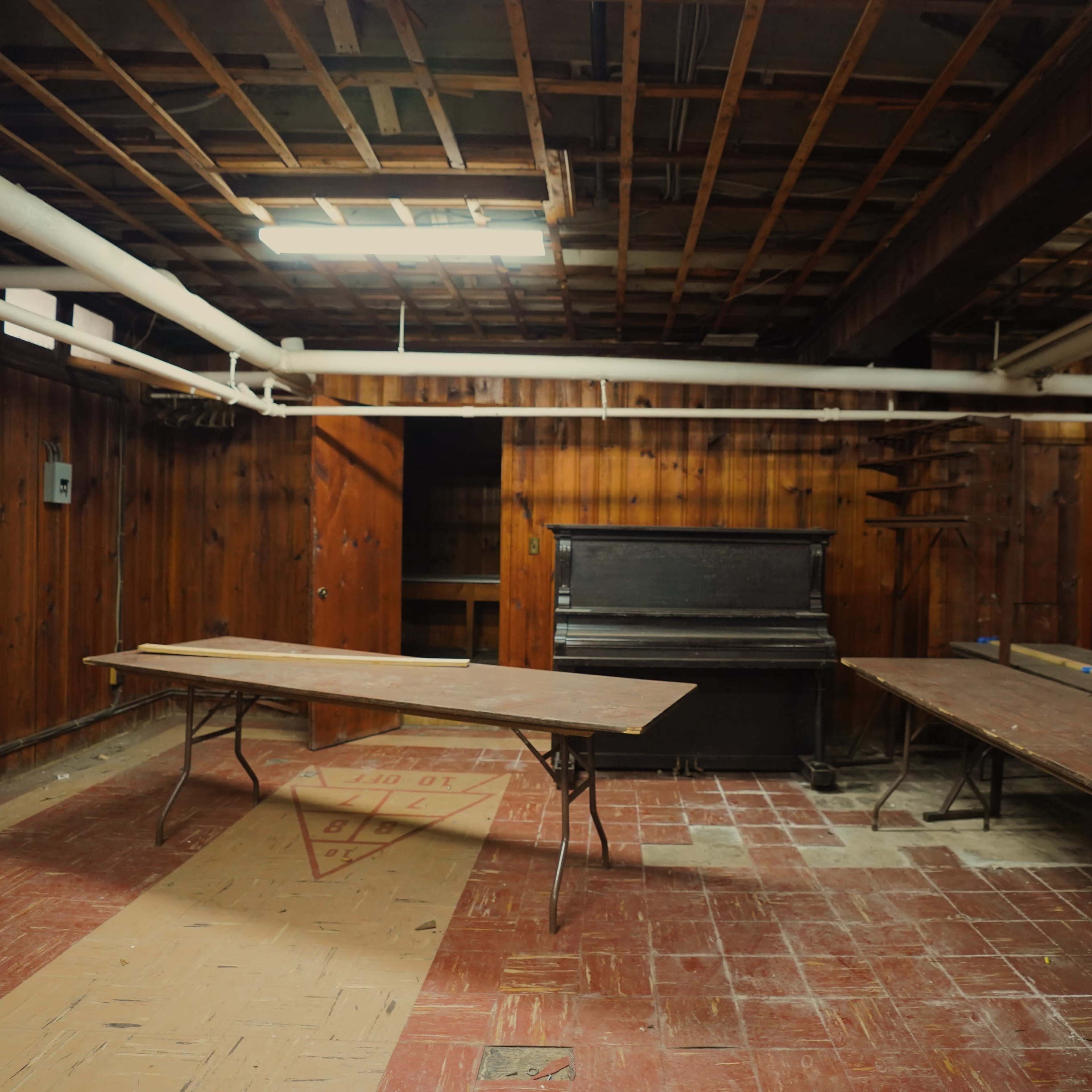 A sparsely furnished basement with wooden paneling, two long tables, and an upright piano.