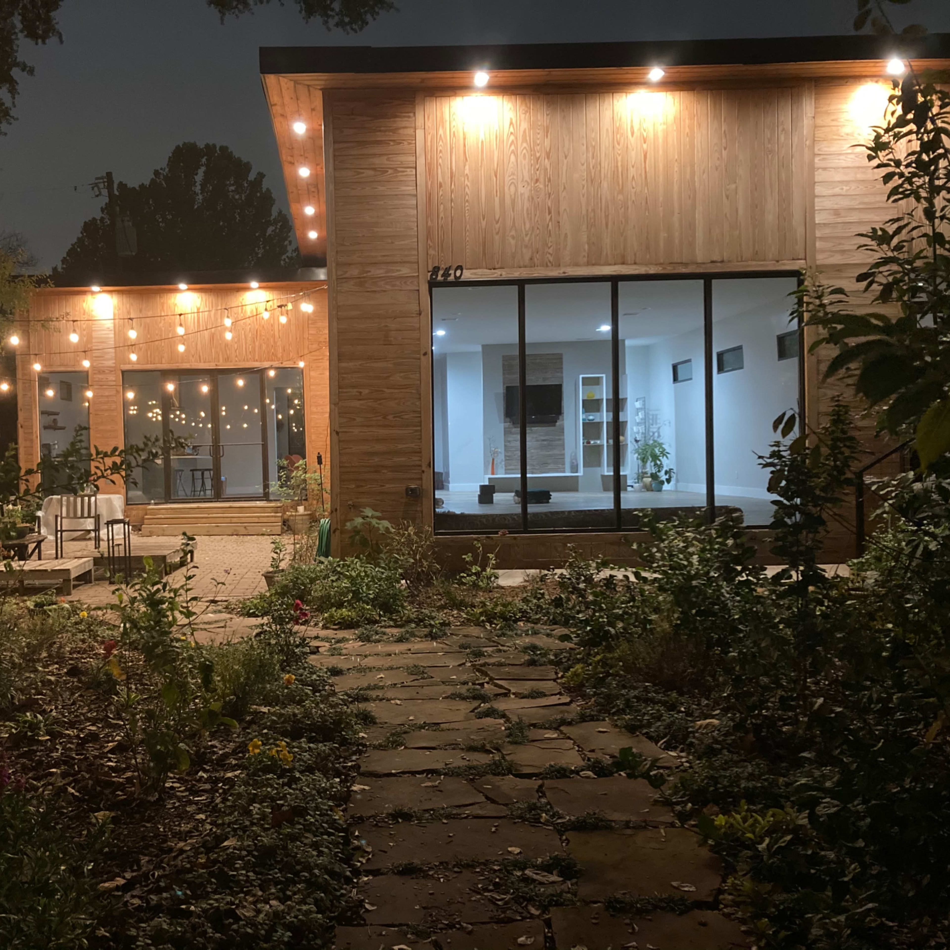 A modern house at night, illuminated by string lights, with a landscaped garden path leading to it.