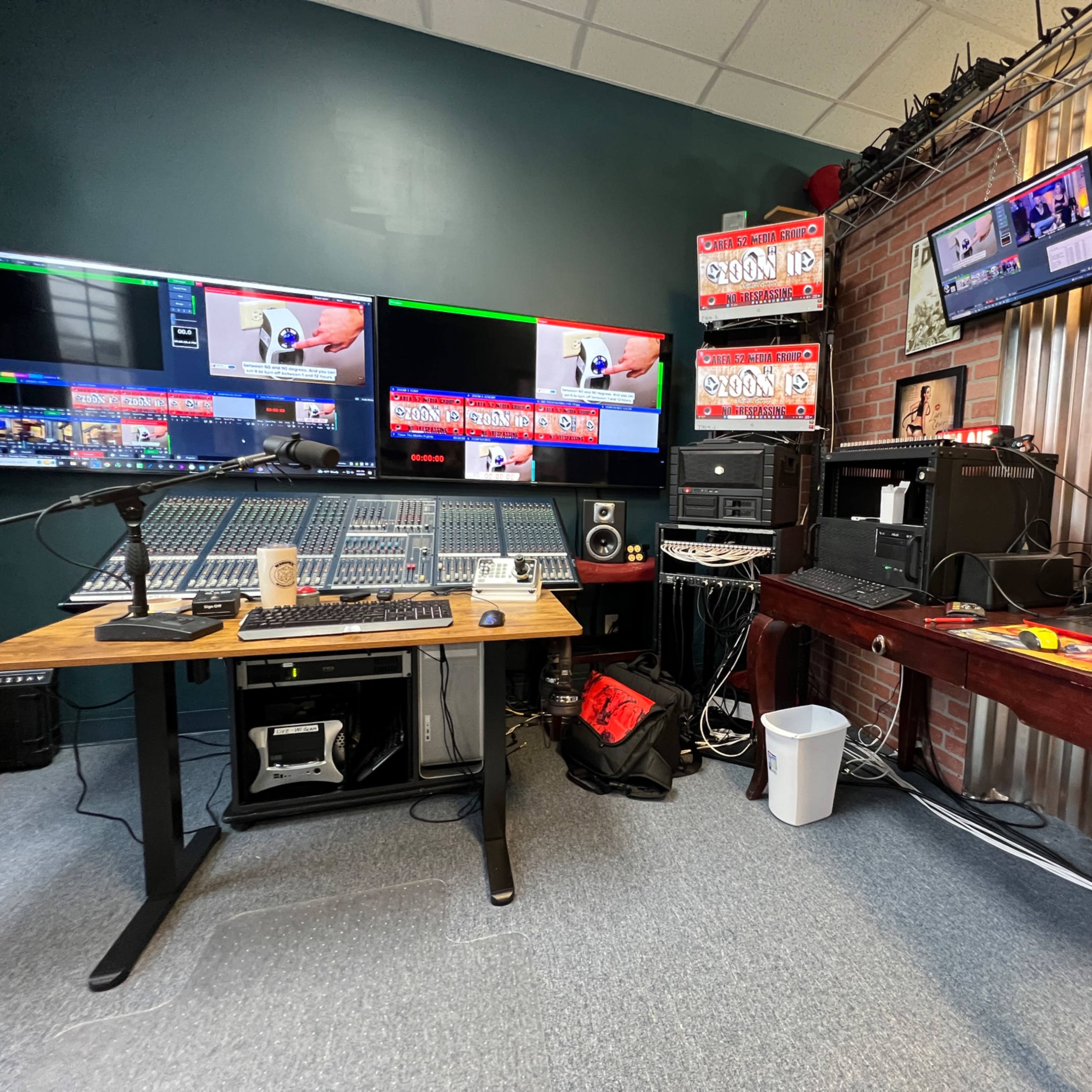 A recording studio with multiple monitors displaying video feeds, a sound mixing console, and various audio equipment arranged around a desk.