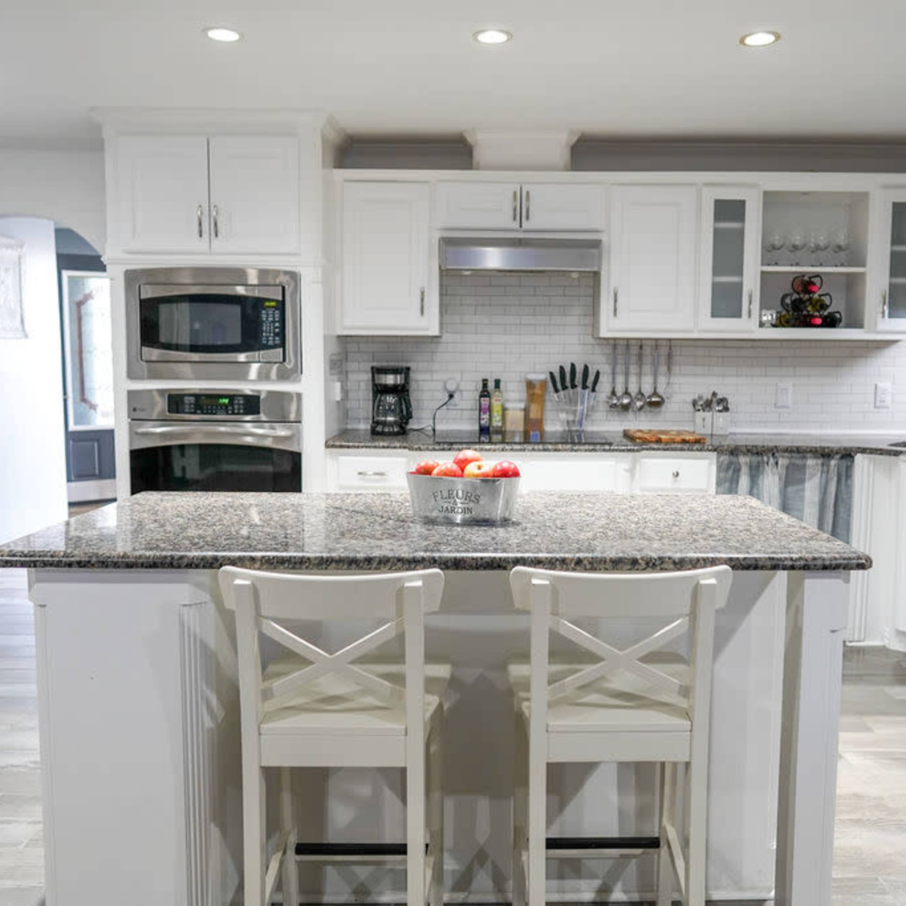 A modern kitchen features a central island with two chairs, white cabinets, stainless steel appliances, and granite countertops.