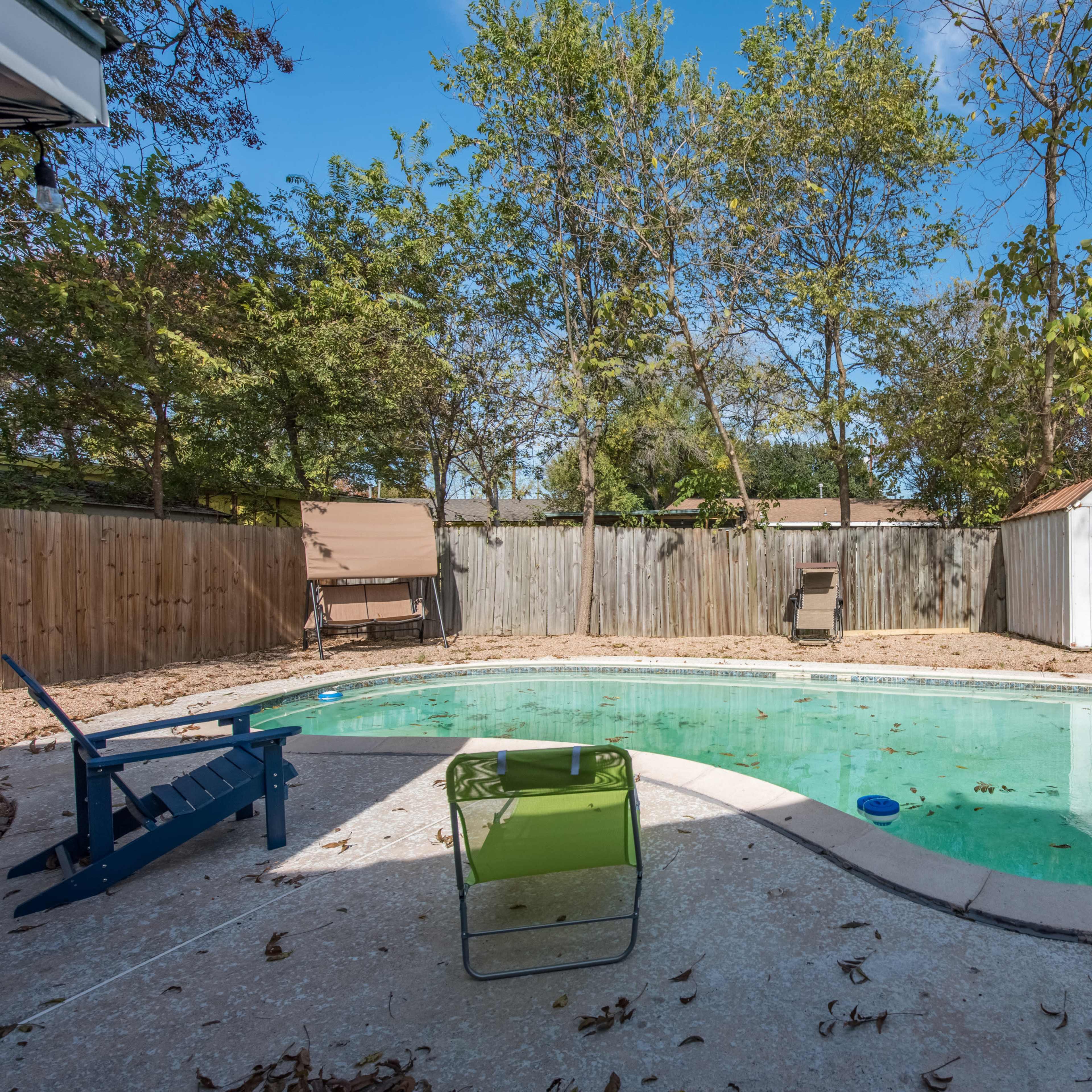 A backyard with a rectangular swimming pool surrounded by a concrete deck, with a wooden fence and a shed in the background.