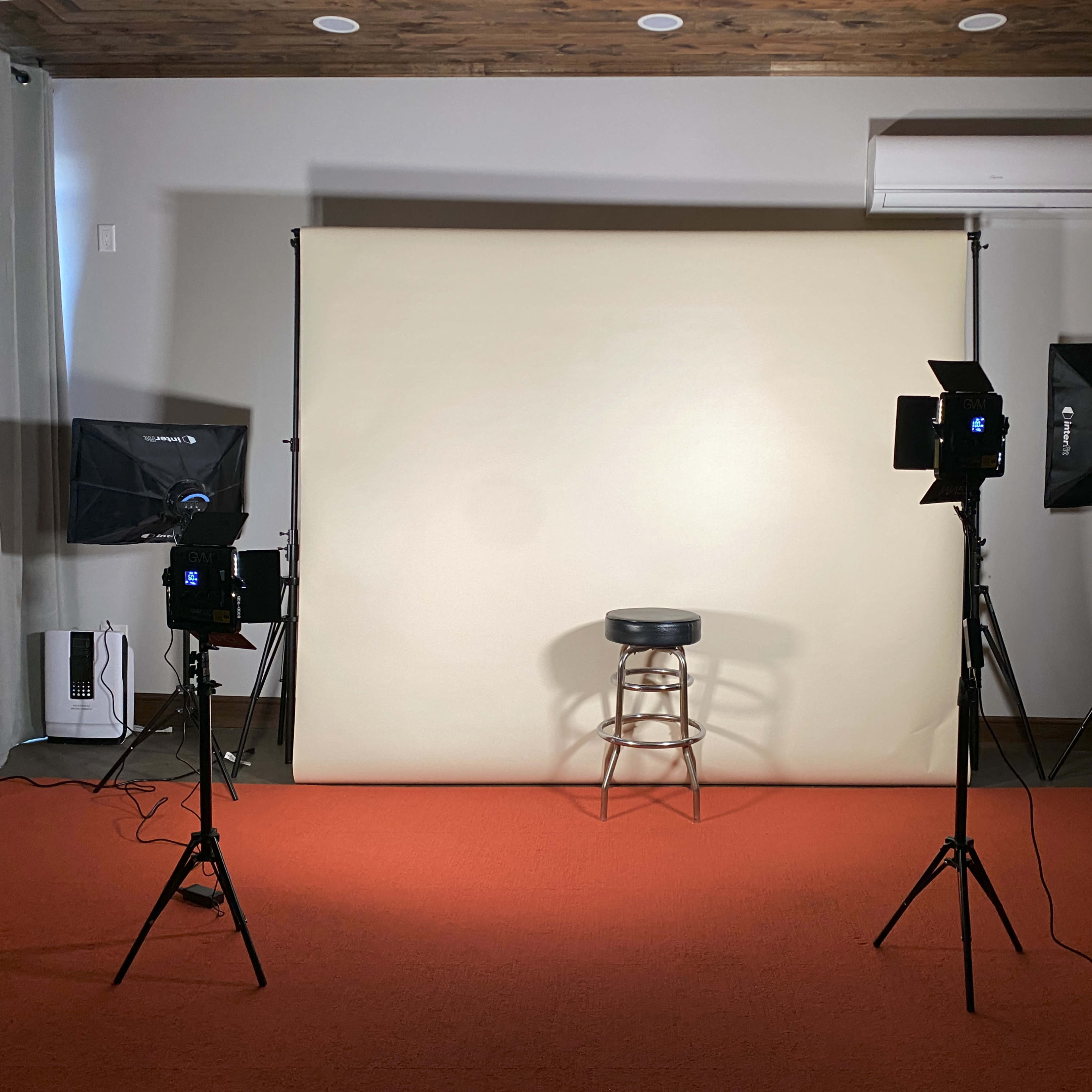 A photo studio setup with a beige backdrop, a black stool in the center, and two lighting stands positioned on either side.
