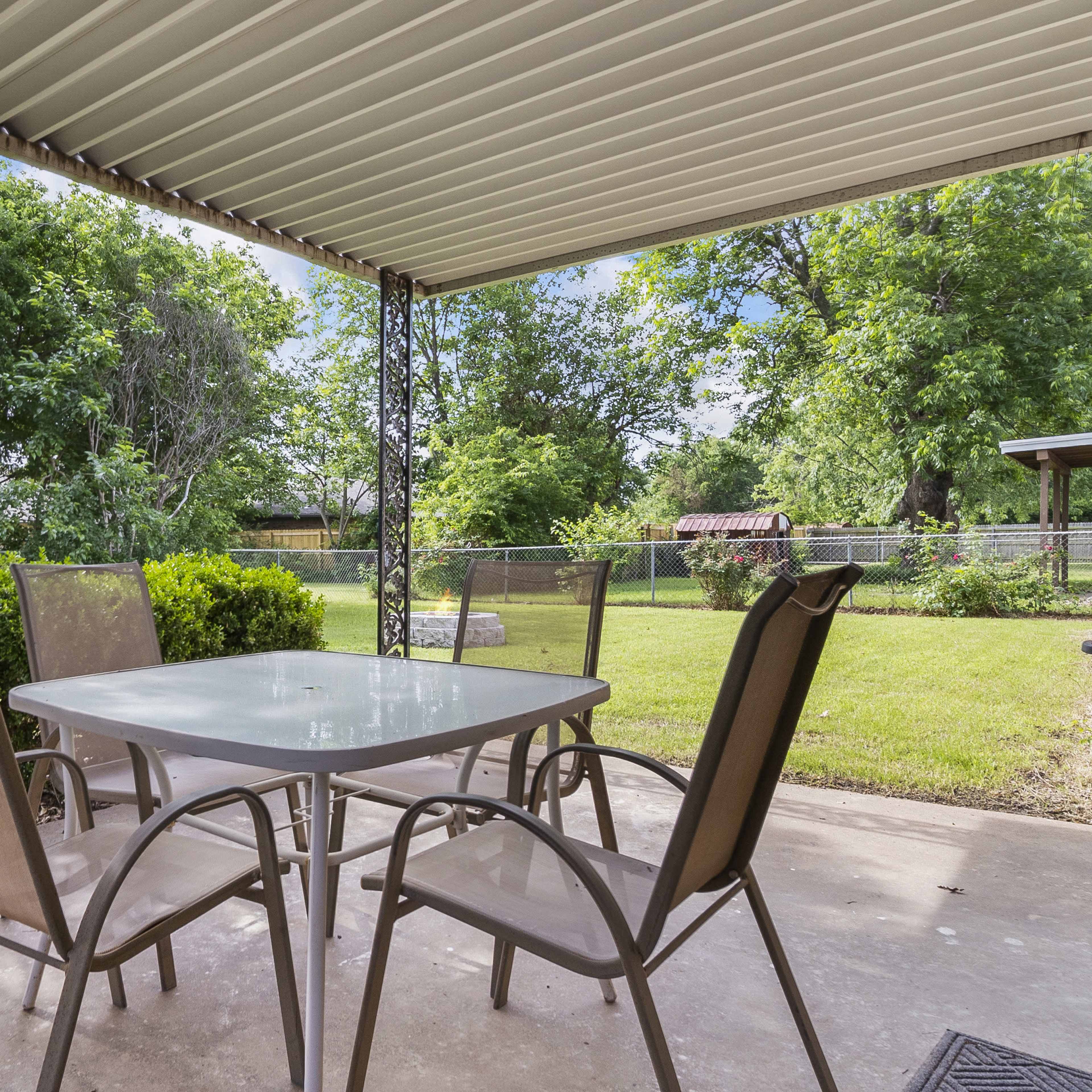 A covered patio features a table with four chairs overlooking a green yard with trees and a fence in the background.