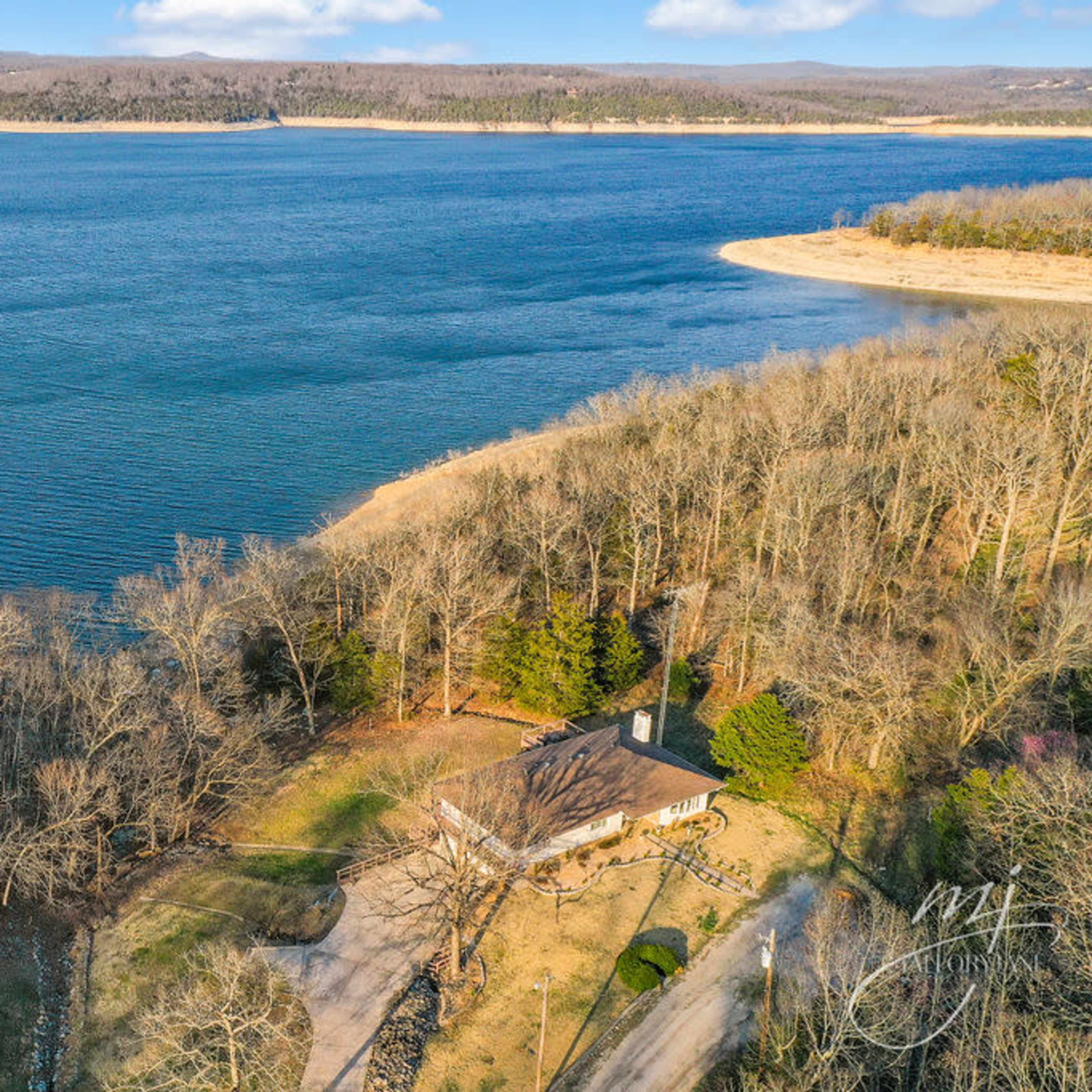 A house situated near the shore of a large lake, surrounded by trees and hills in the background.