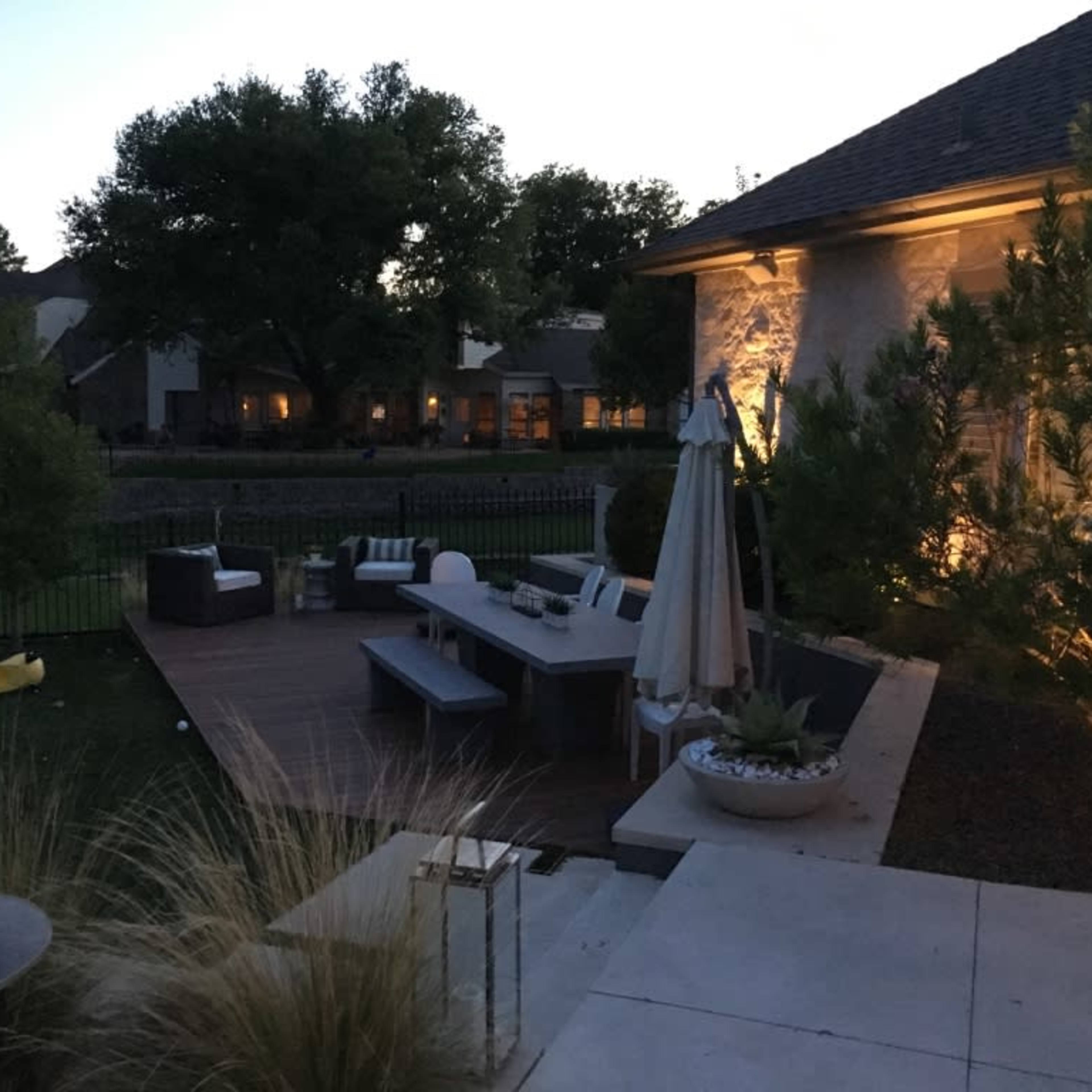 A landscaped backyard at dusk, featuring a wooden deck with outdoor furniture, a table set for dining, and a pool area illuminated by soft lighting.