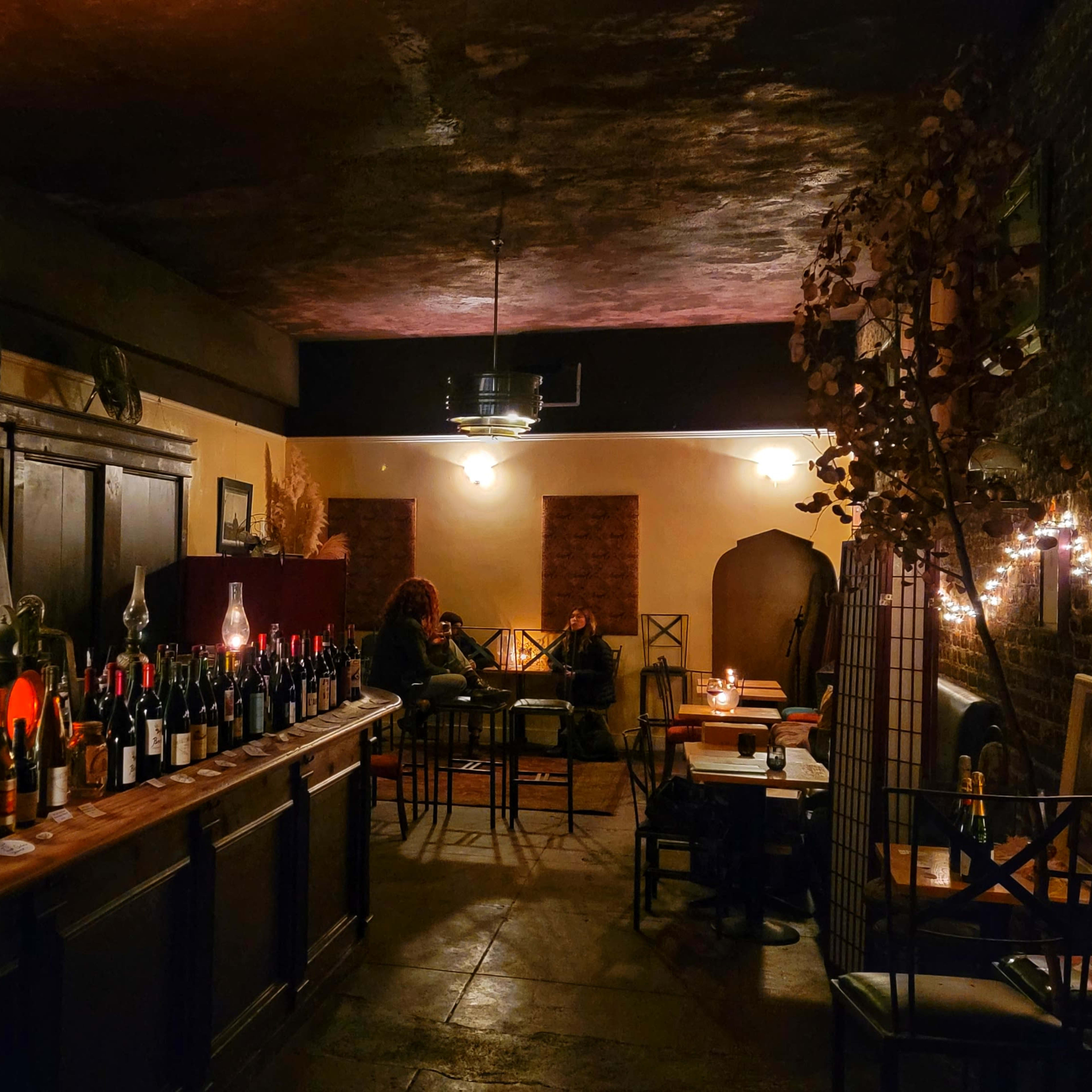 A dimly lit bar area features a long wooden counter with an array of bottles and tables in the background.