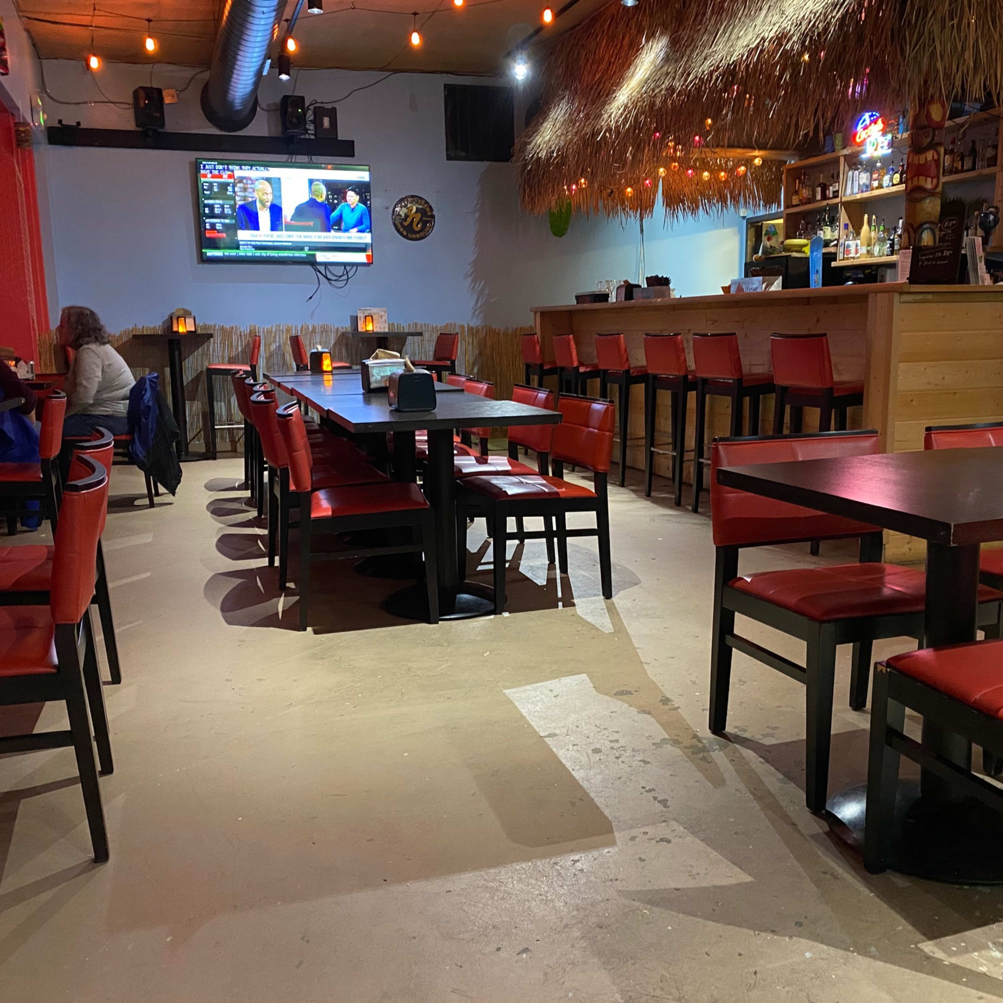An empty restaurant interior featuring black tables and red chairs, with a bar area and multiple televisions displaying content.