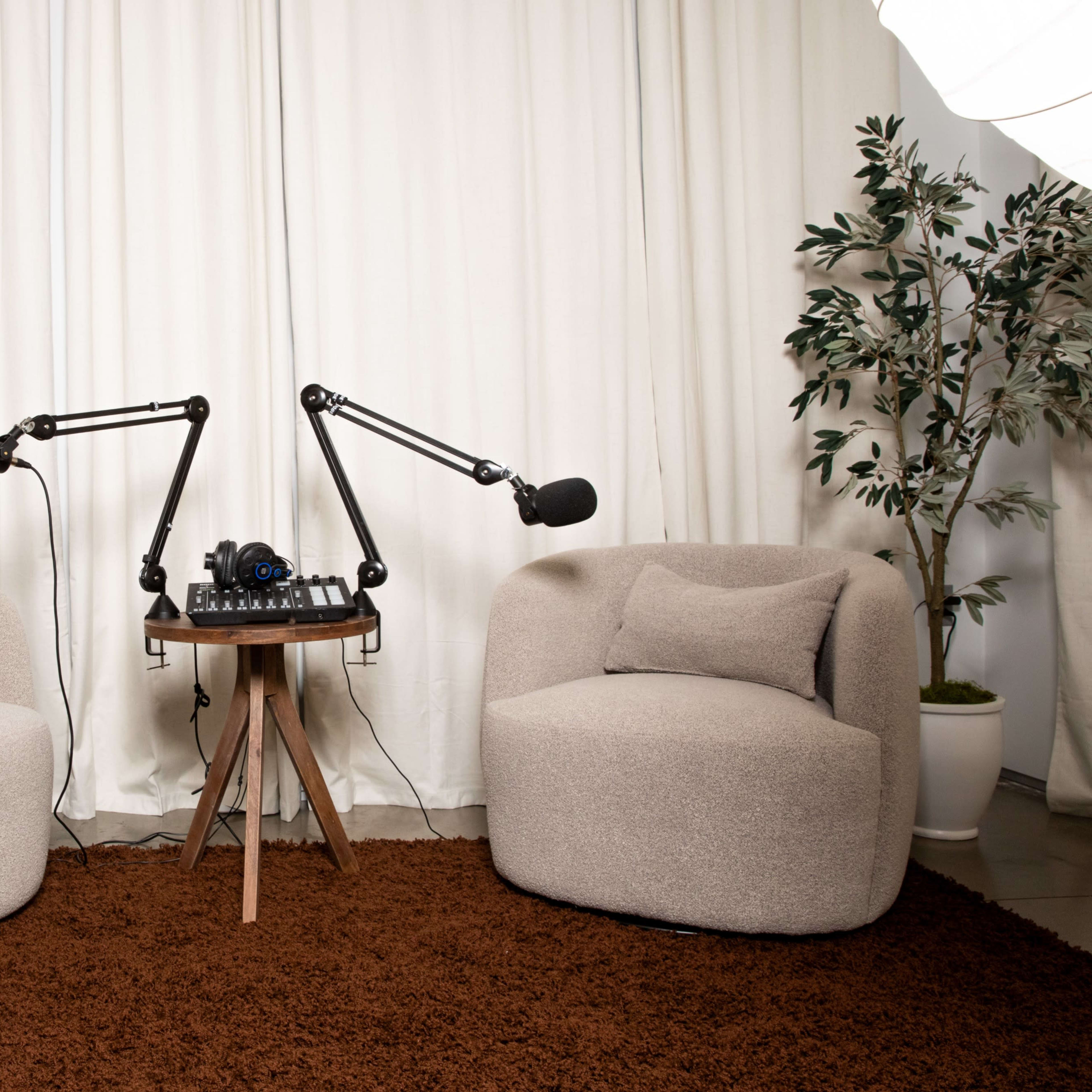 Two upholstered chairs facing a small table equipped with microphones and audio equipment, set against a backdrop of a white curtain and a plant.
