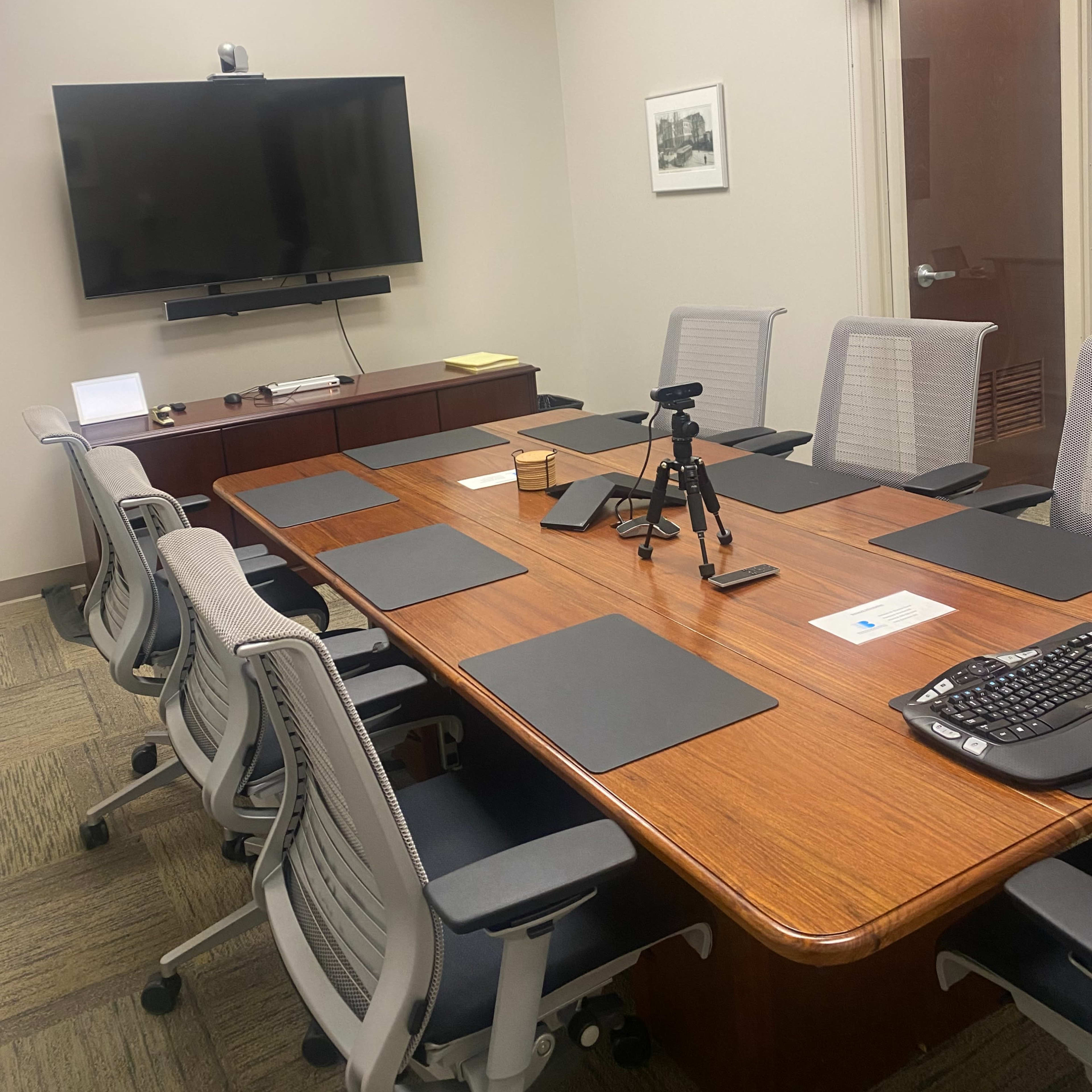 A conference room with a large wooden table surrounded by ergonomic chairs, equipped with a TV on the wall and a tripod on the table.