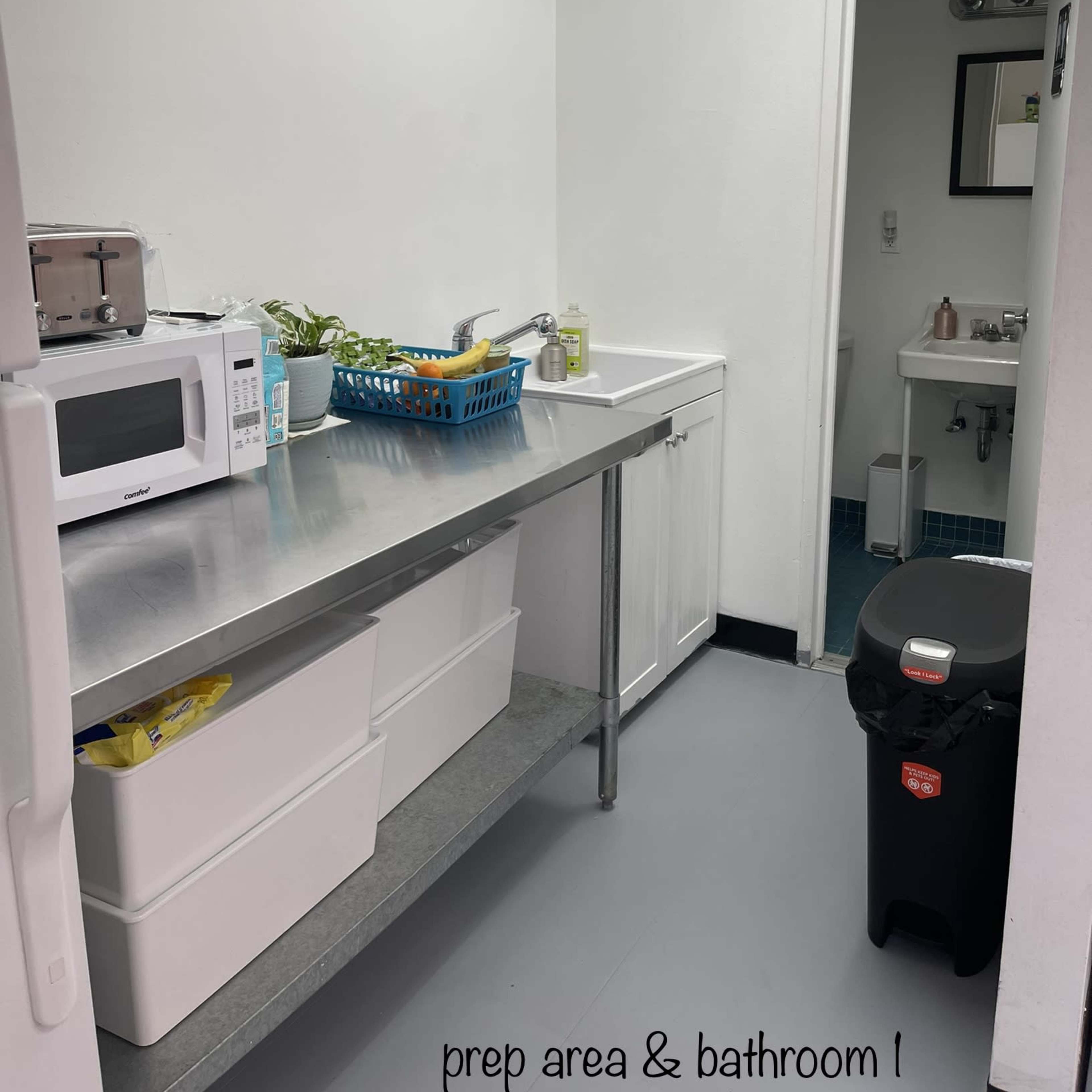 A kitchen prep area with a stainless steel table, a microwave, a small plant, and a trash can, alongside a bathroom visible through an open door.