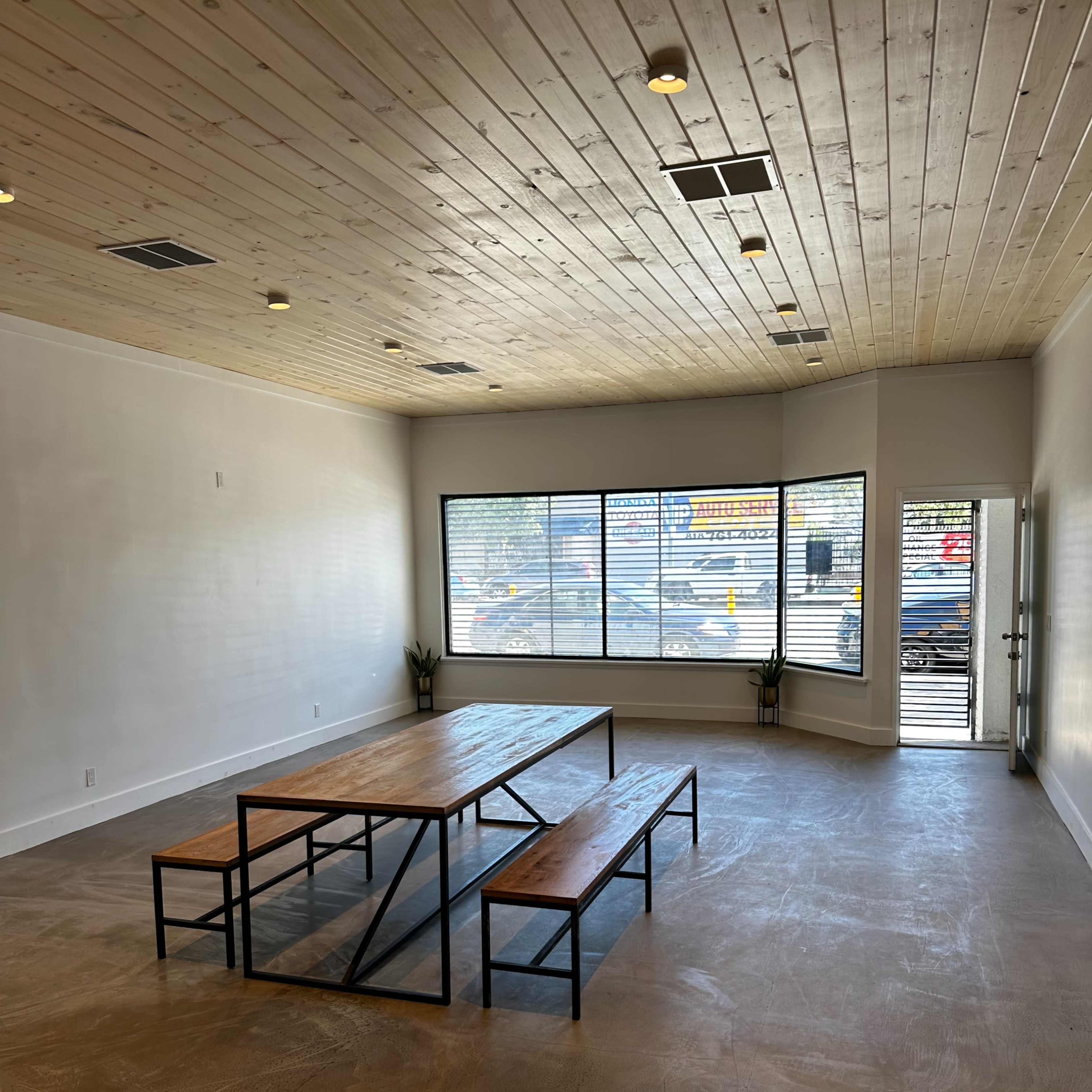 A spacious, well-lit interior featuring a wooden table with two benches and large windows allowing natural light to enter.