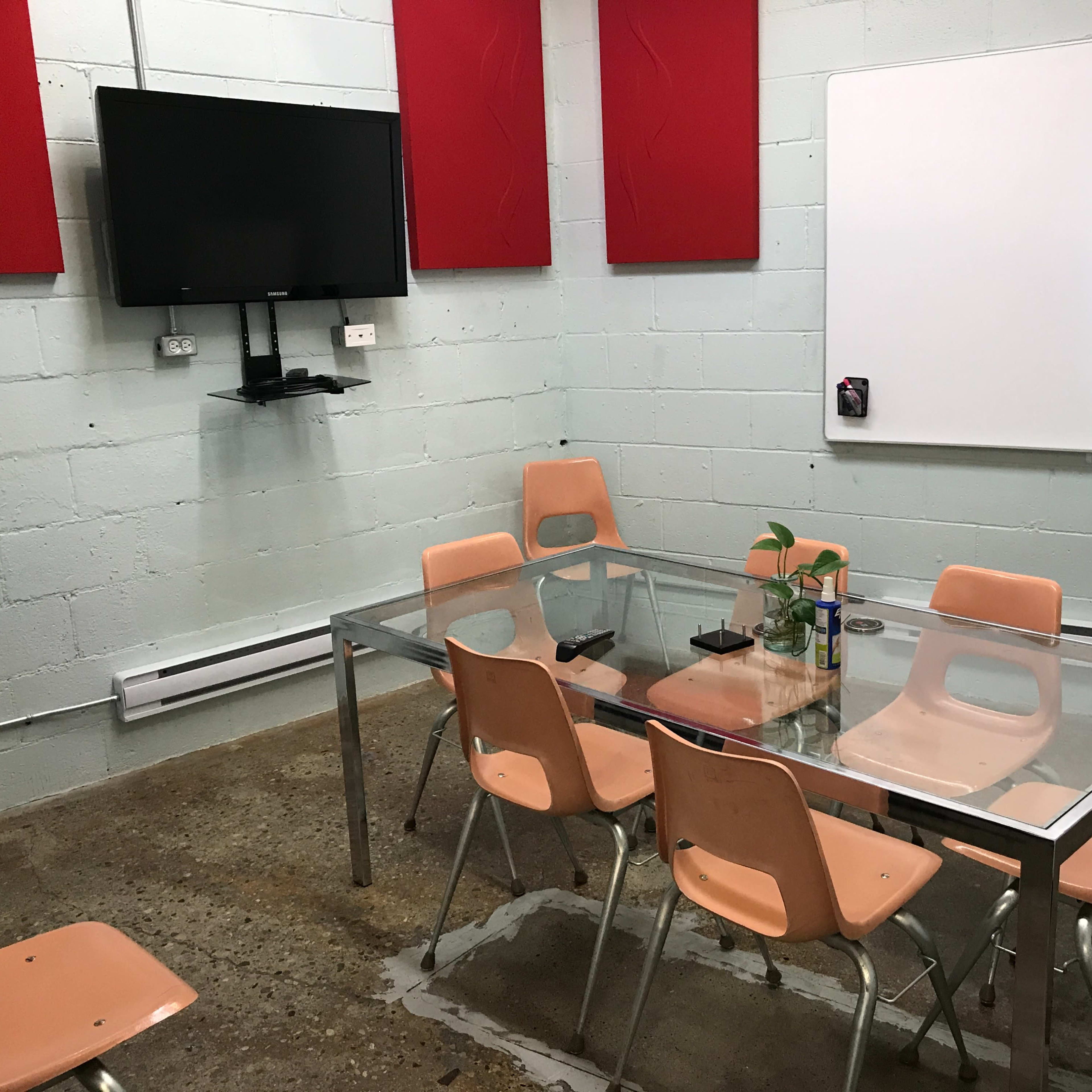 A meeting room features a glass table surrounded by eight peach-colored chairs, a wall-mounted television, and a whiteboard.