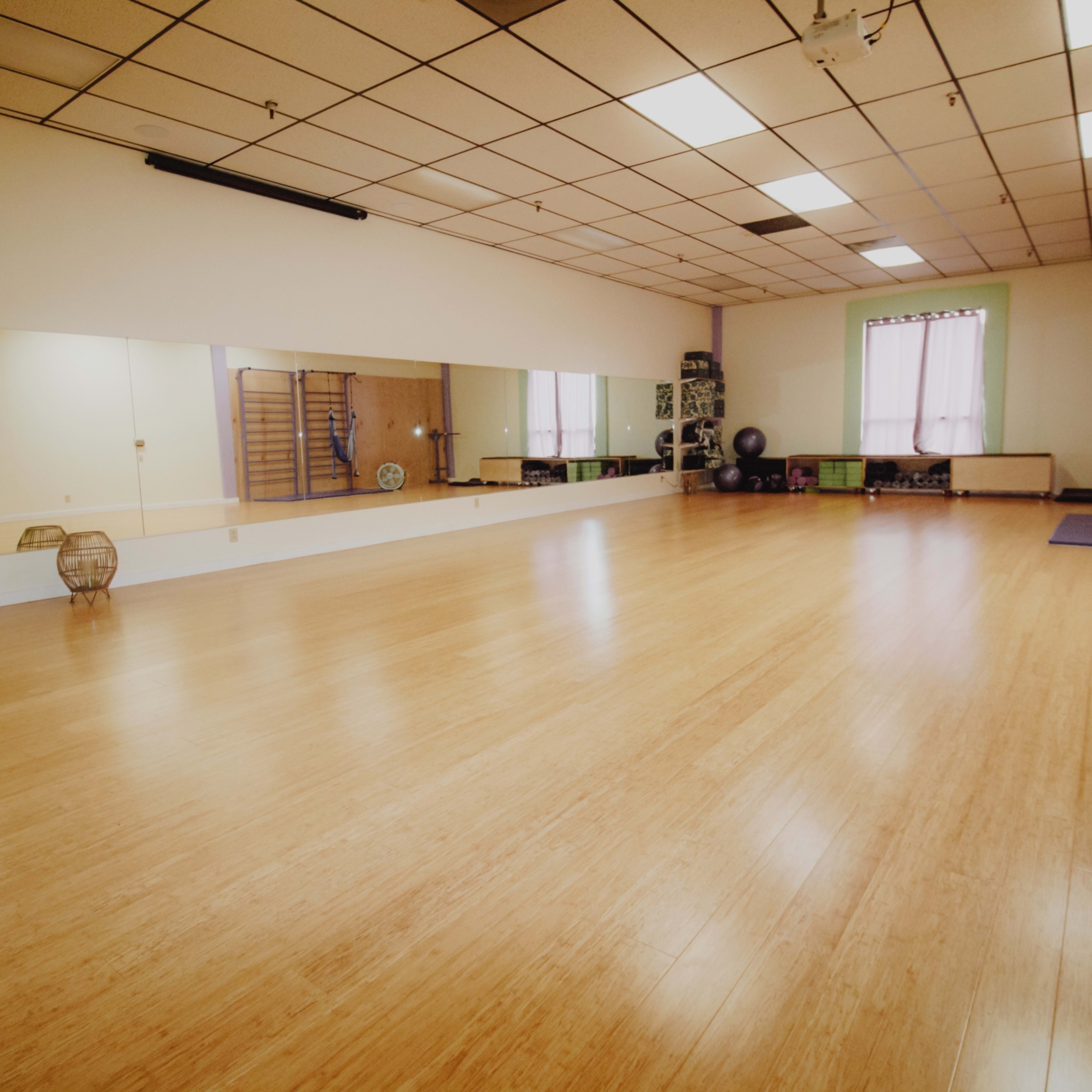 An empty fitness studio with wooden flooring, mirrors along one wall, and equipment and fans in the corners.