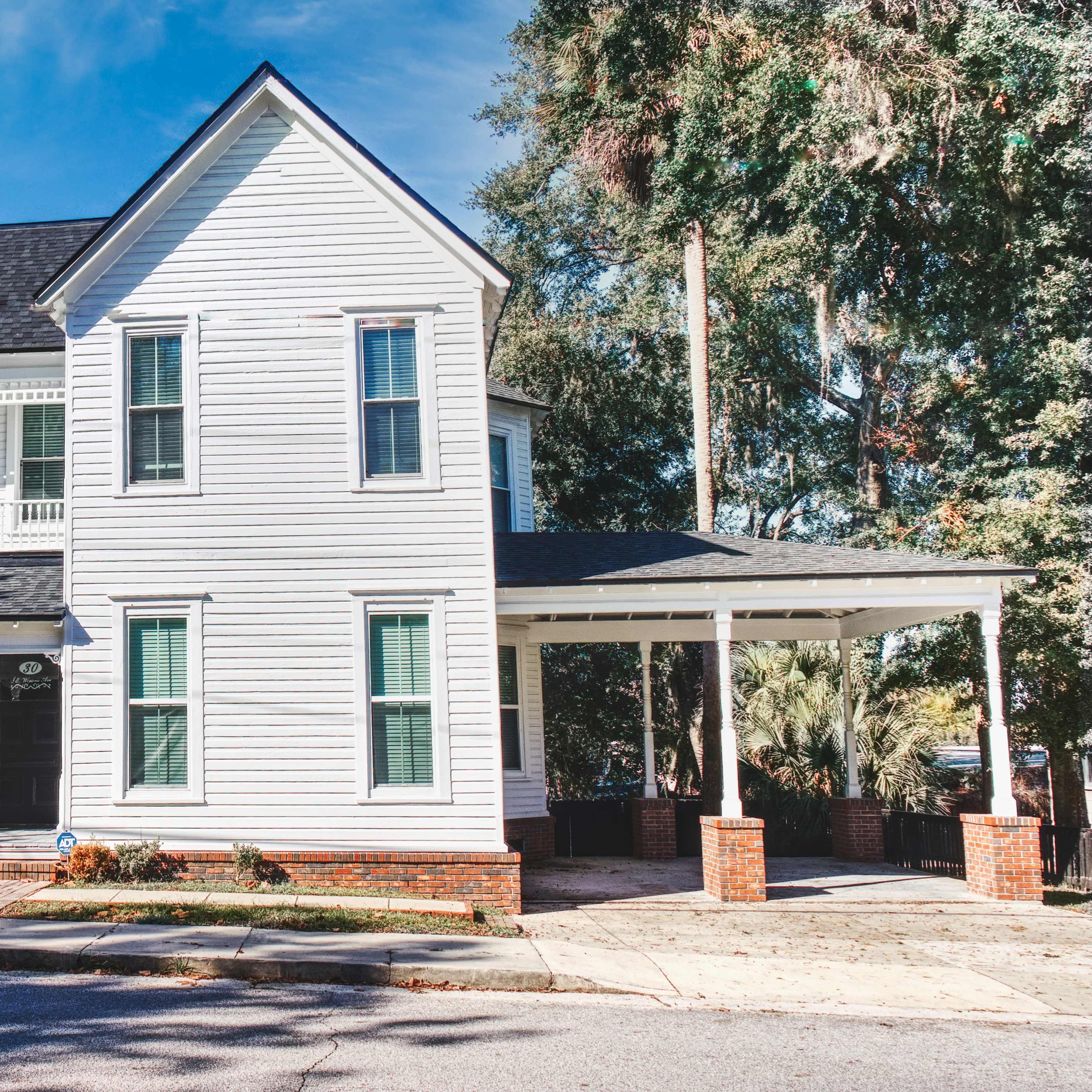 A two-story white wooden house with a black roof and a covered porch is situated on a street corner.