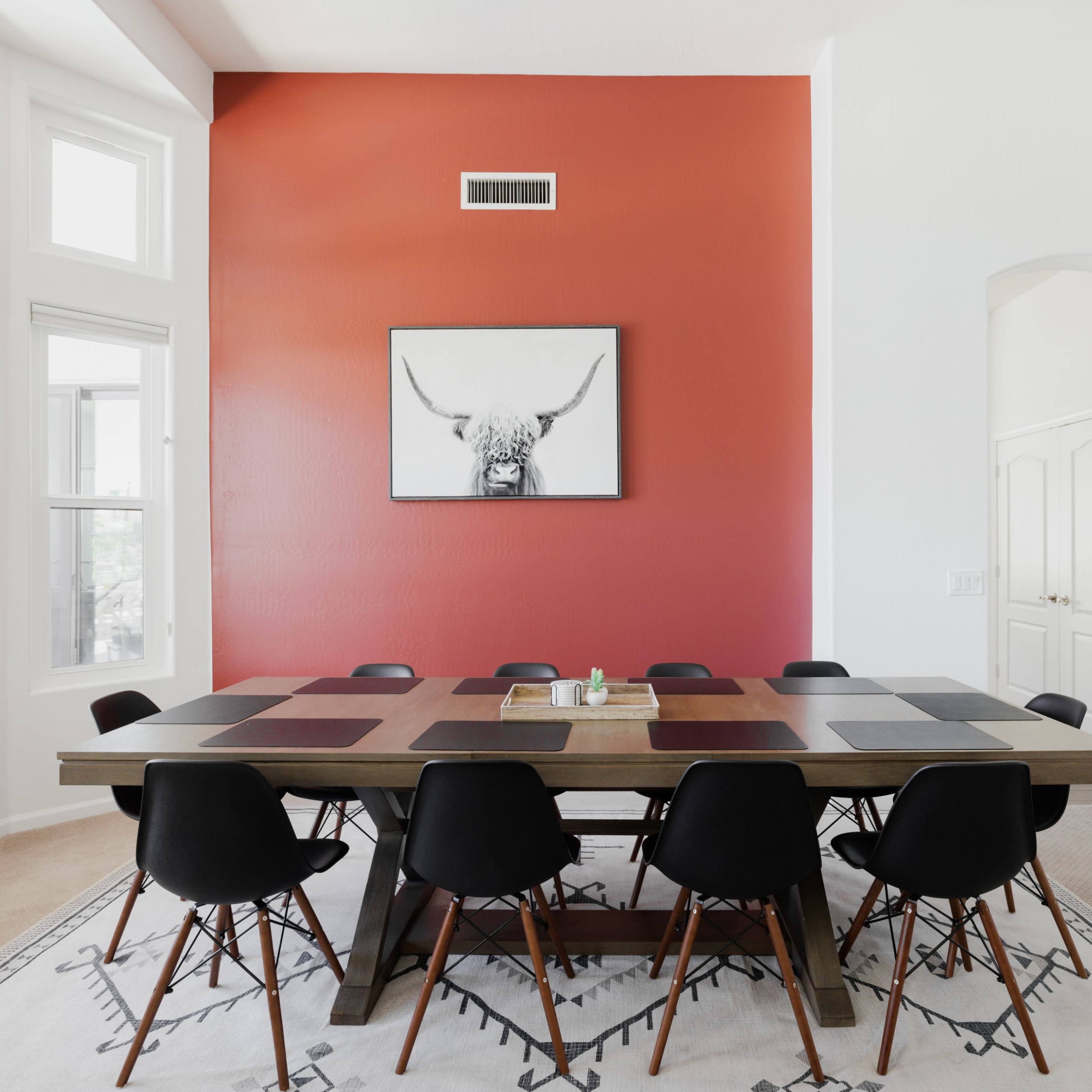 A long wooden dining table with black chairs is set against a red accent wall featuring a framed picture of a bull's head.