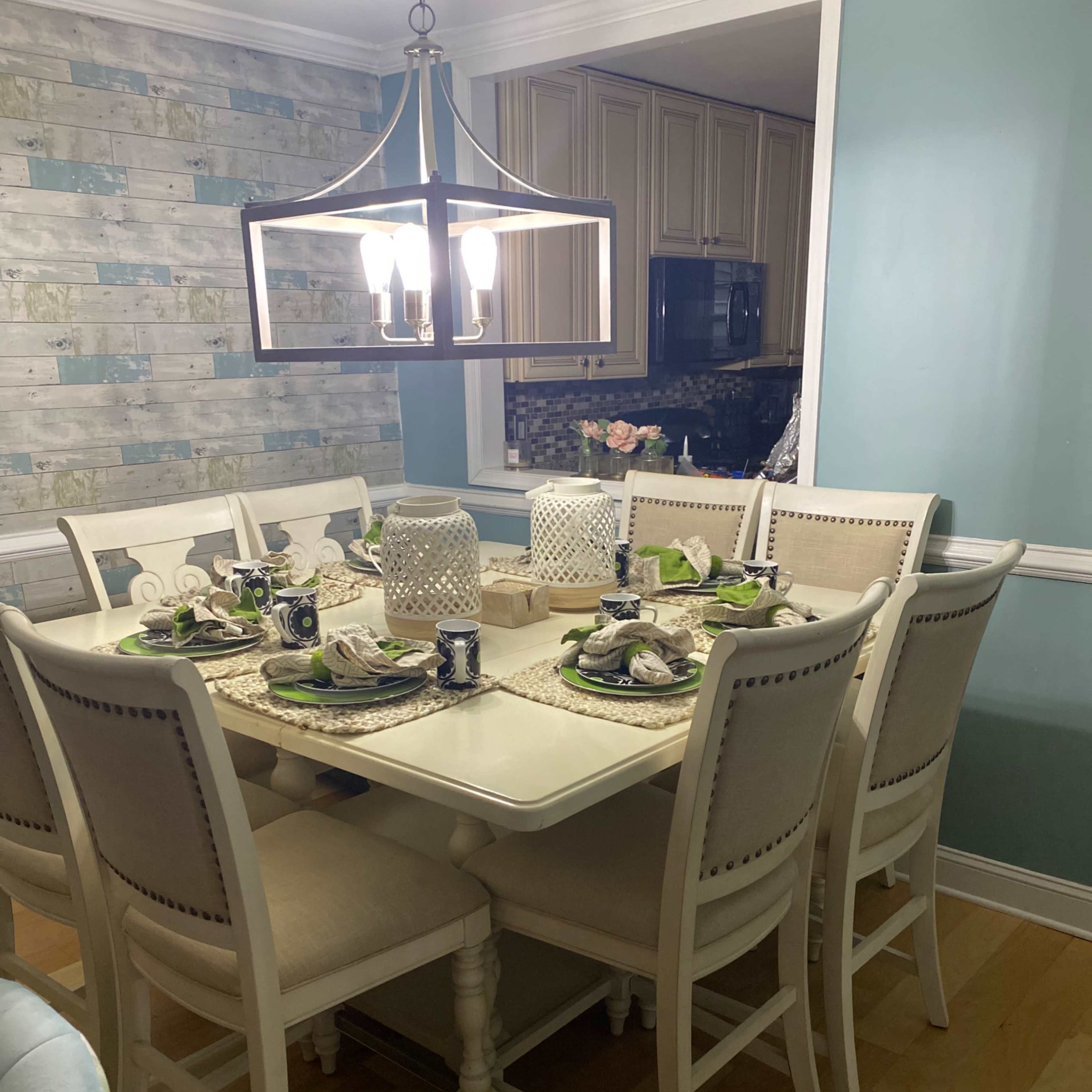 A light-colored dining room table is set with plates, glasses, and decorative centerpieces, surrounded by eight upholstered chairs.