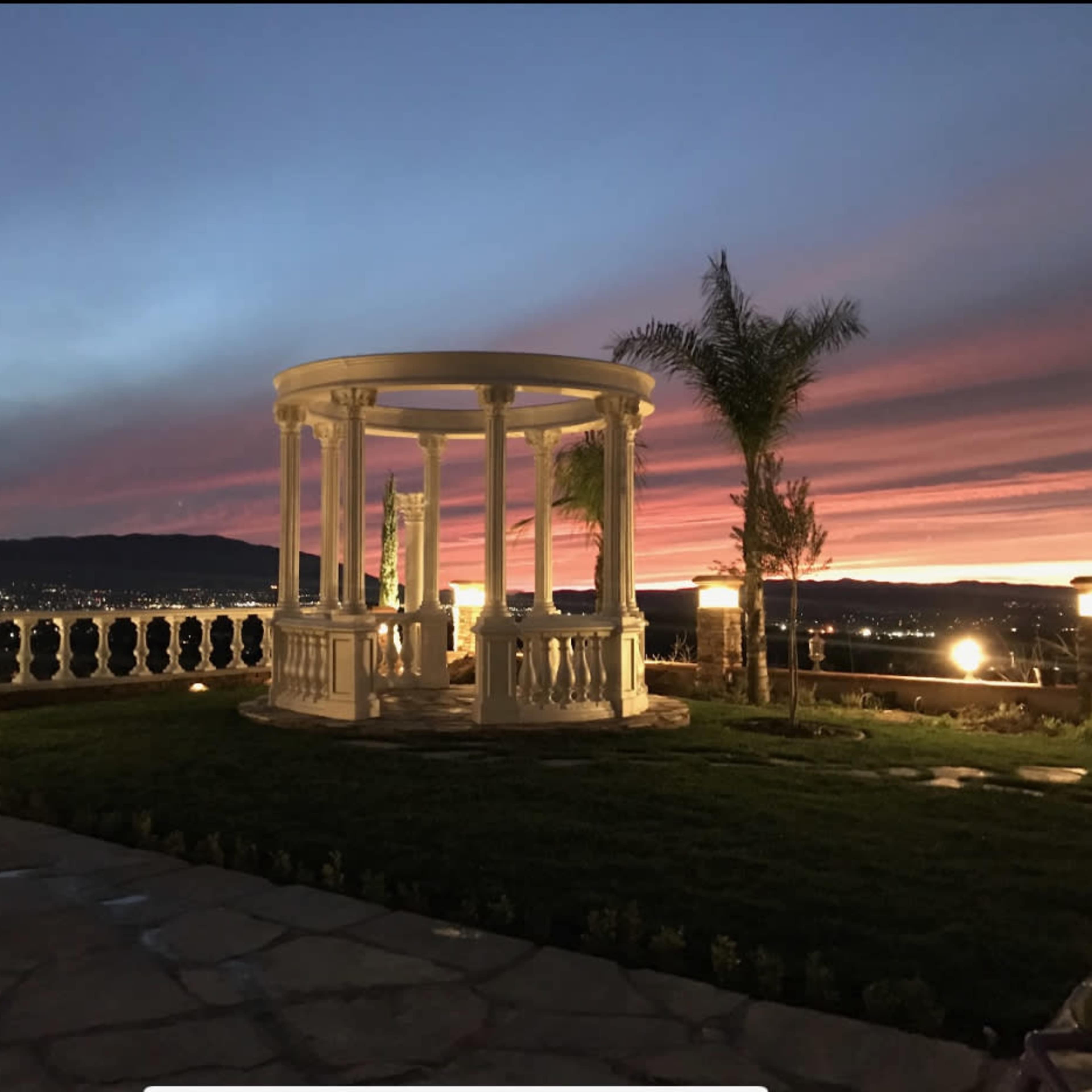 A white gazebo with columns is situated on a landscaped terrace, overlooking a valley at sunset with colorful clouds.