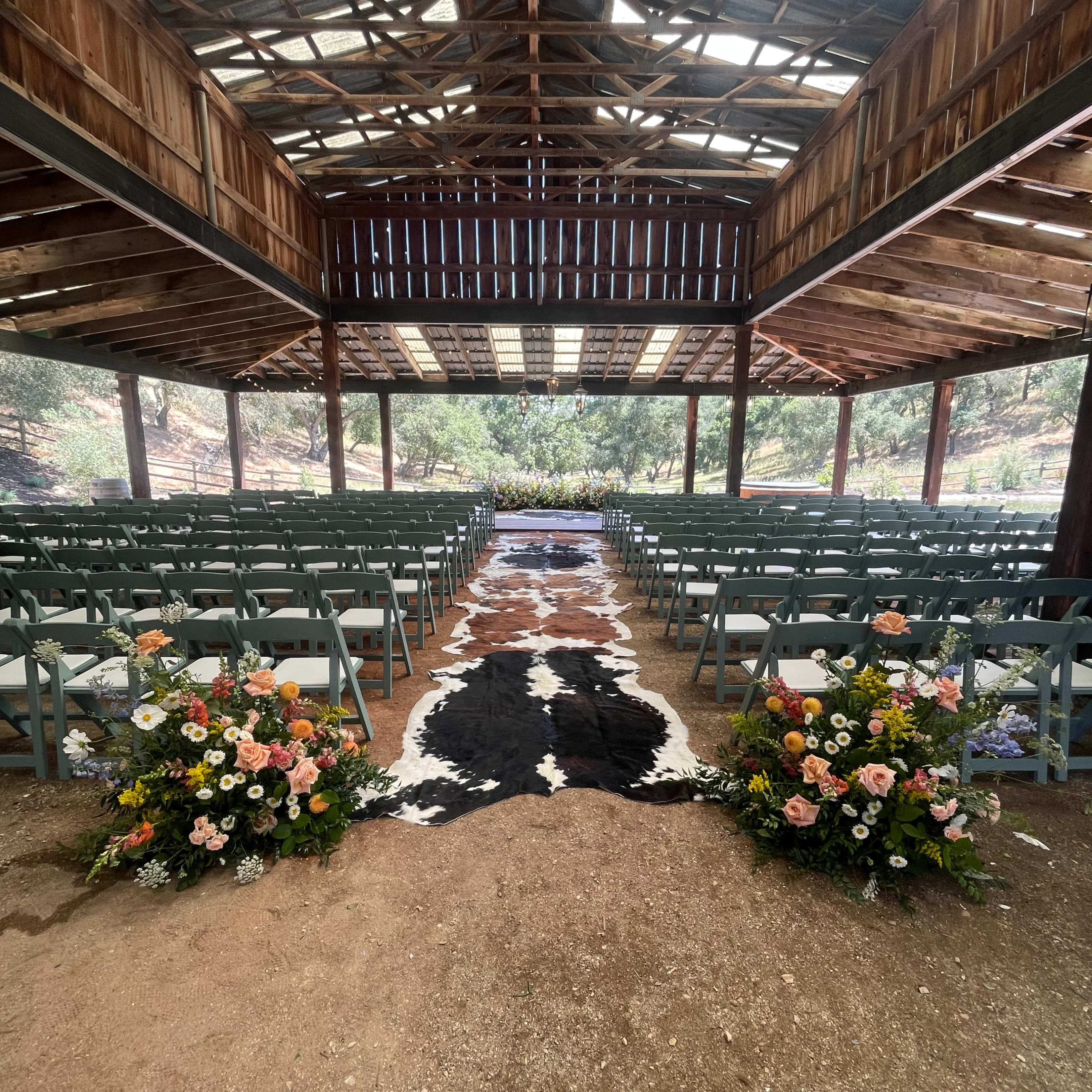 A rustic outdoor venue features rows of chairs facing a central aisle adorned with a cowhide rug and floral arrangements on either side.