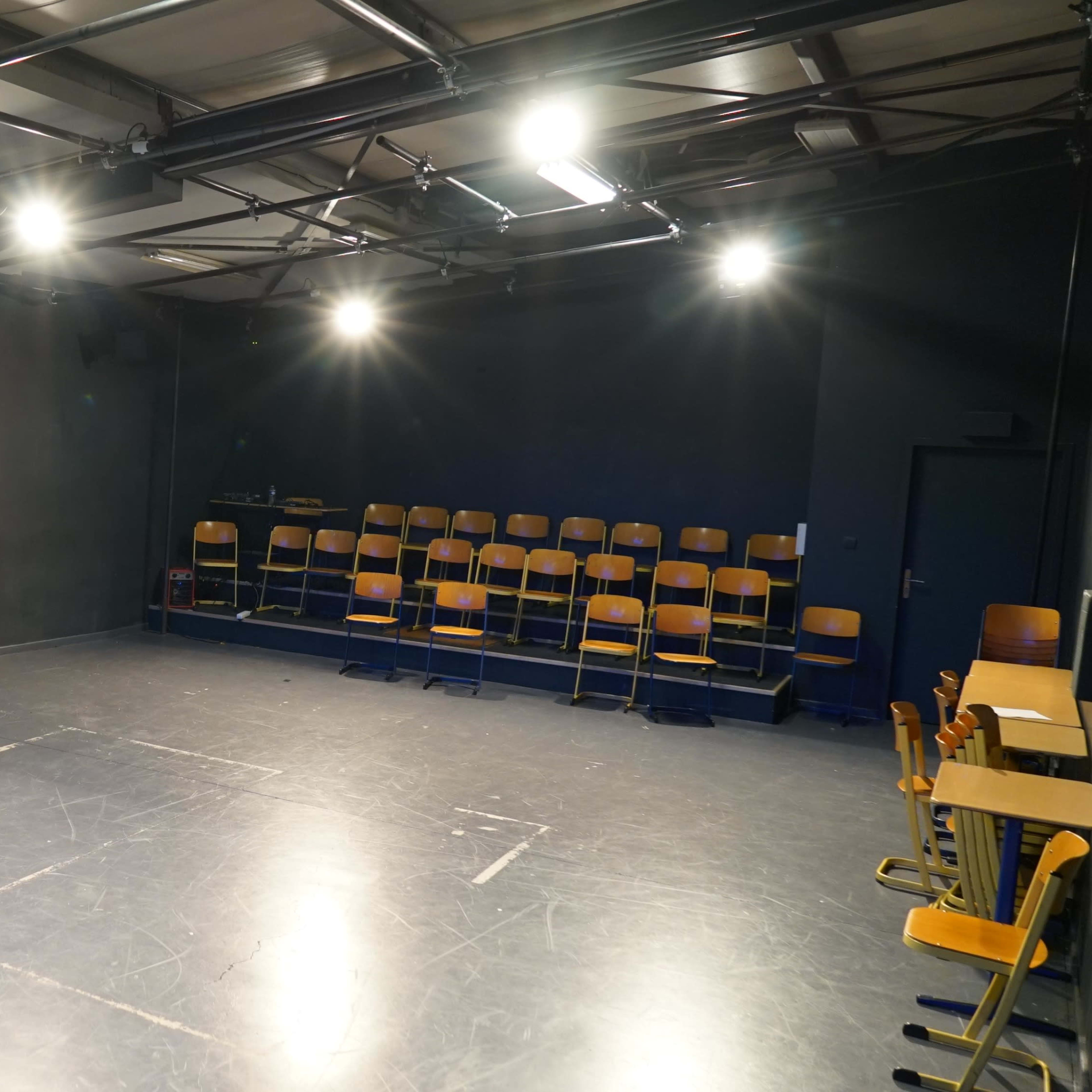 A sparsely furnished theater stage with a dark backdrop, illuminated by overhead lights and featuring rows of wooden chairs against one wall.