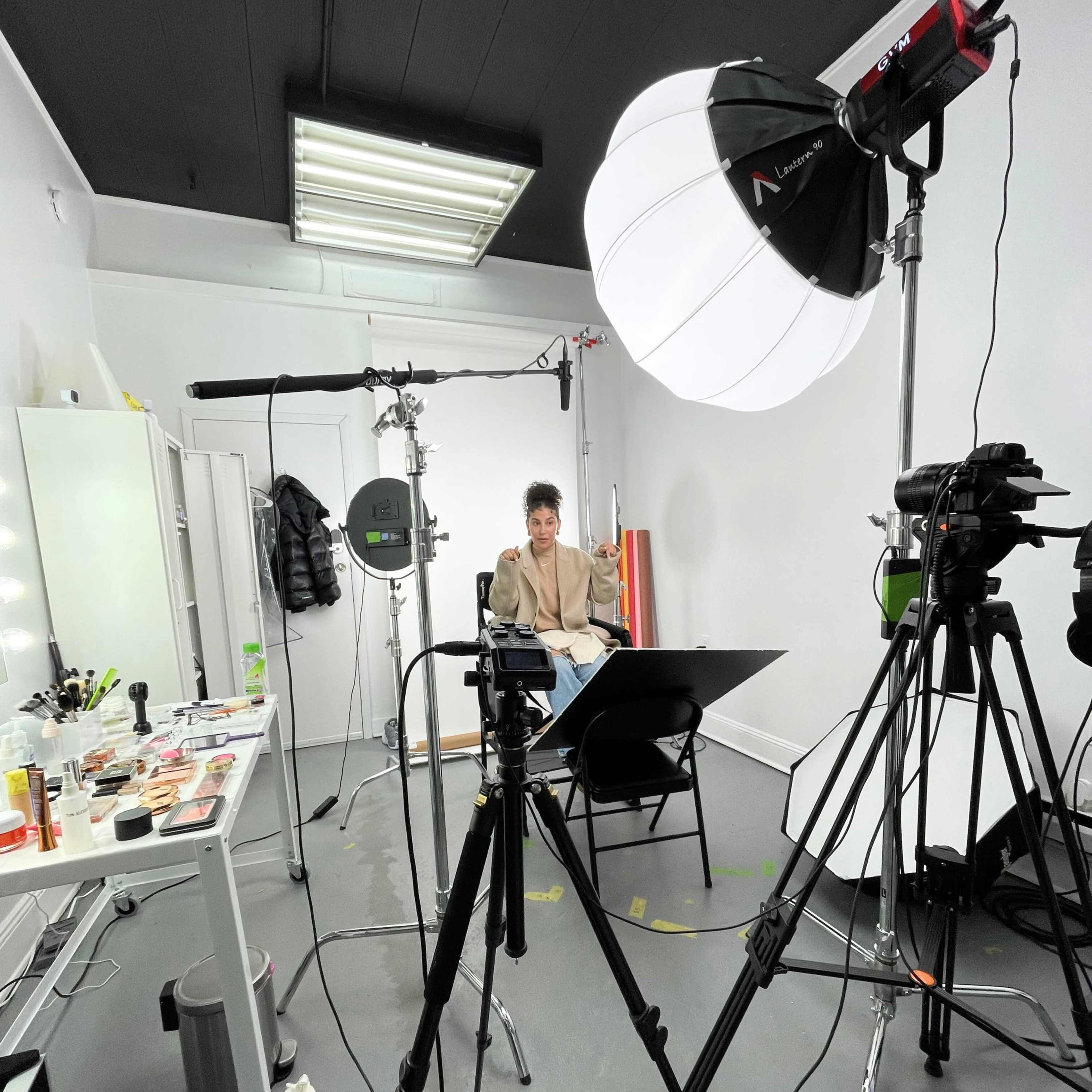 A studio setup features a person seated in front of cameras and lighting equipment with a table displaying various makeup products.
