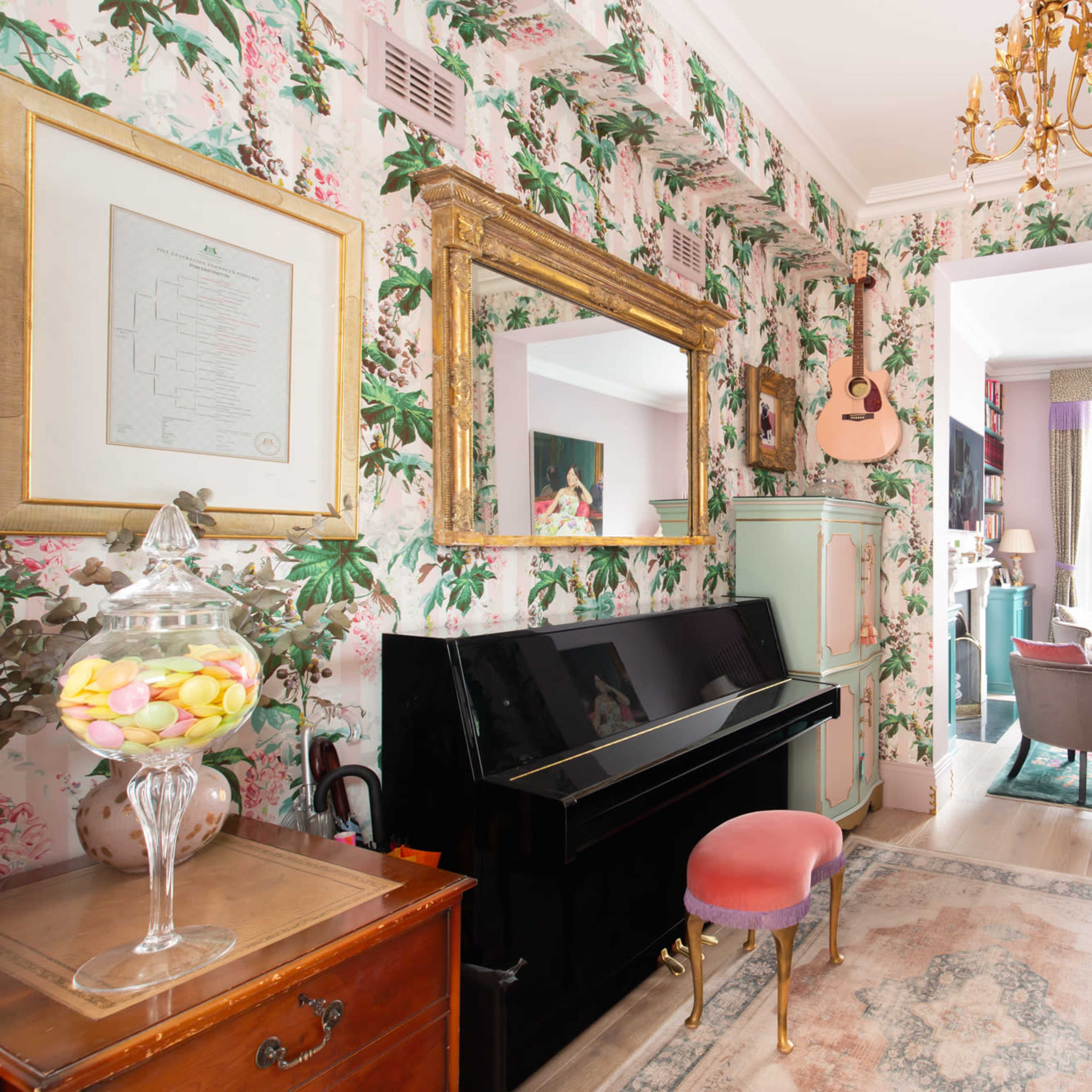 A narrow hallway with floral wallpaper, featuring a black piano, a vintage wooden cabinet, and a doorway leading to a brightly lit living area.