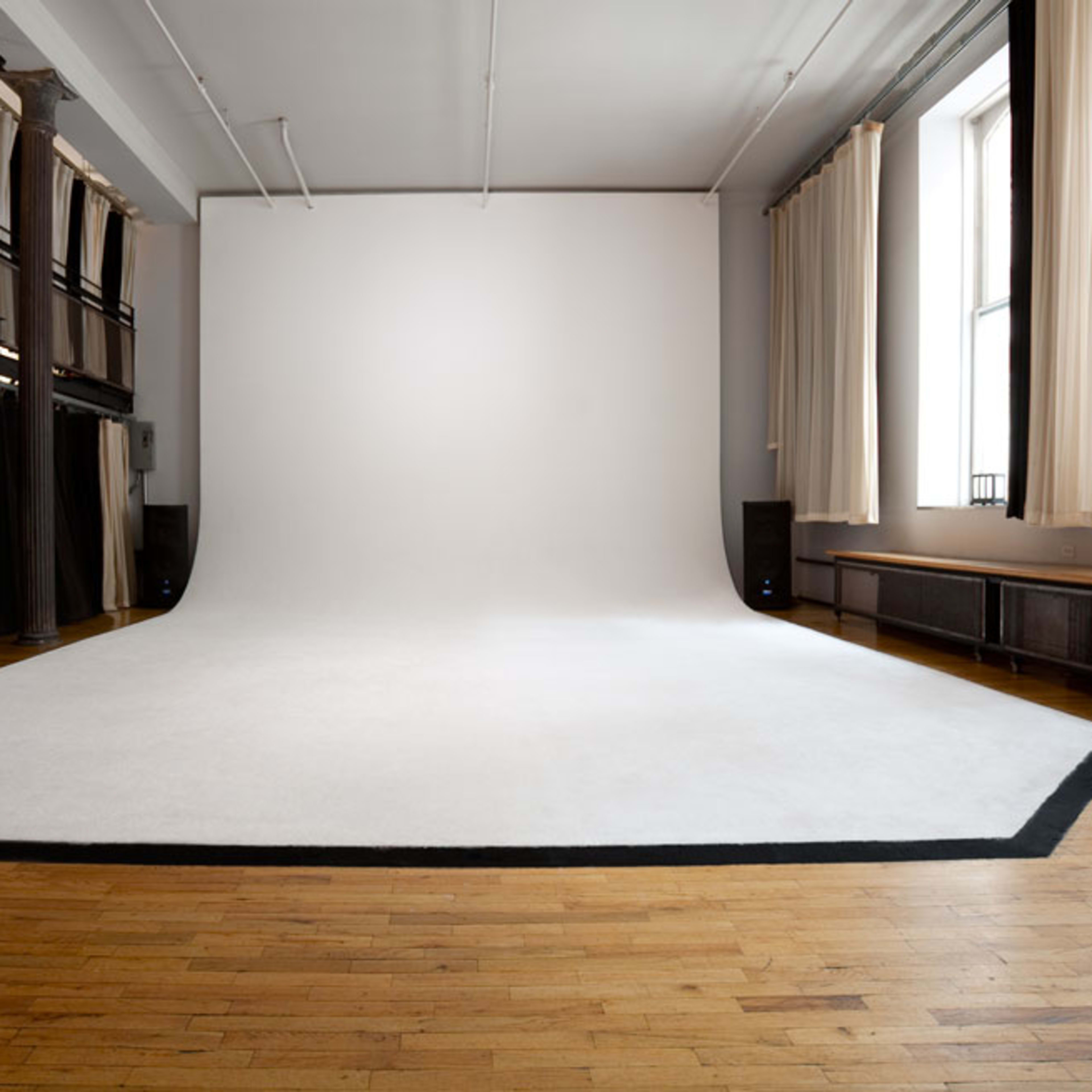 A spacious photo studio with a large, white backdrop and a wooden floor, illuminated by natural light from windows.