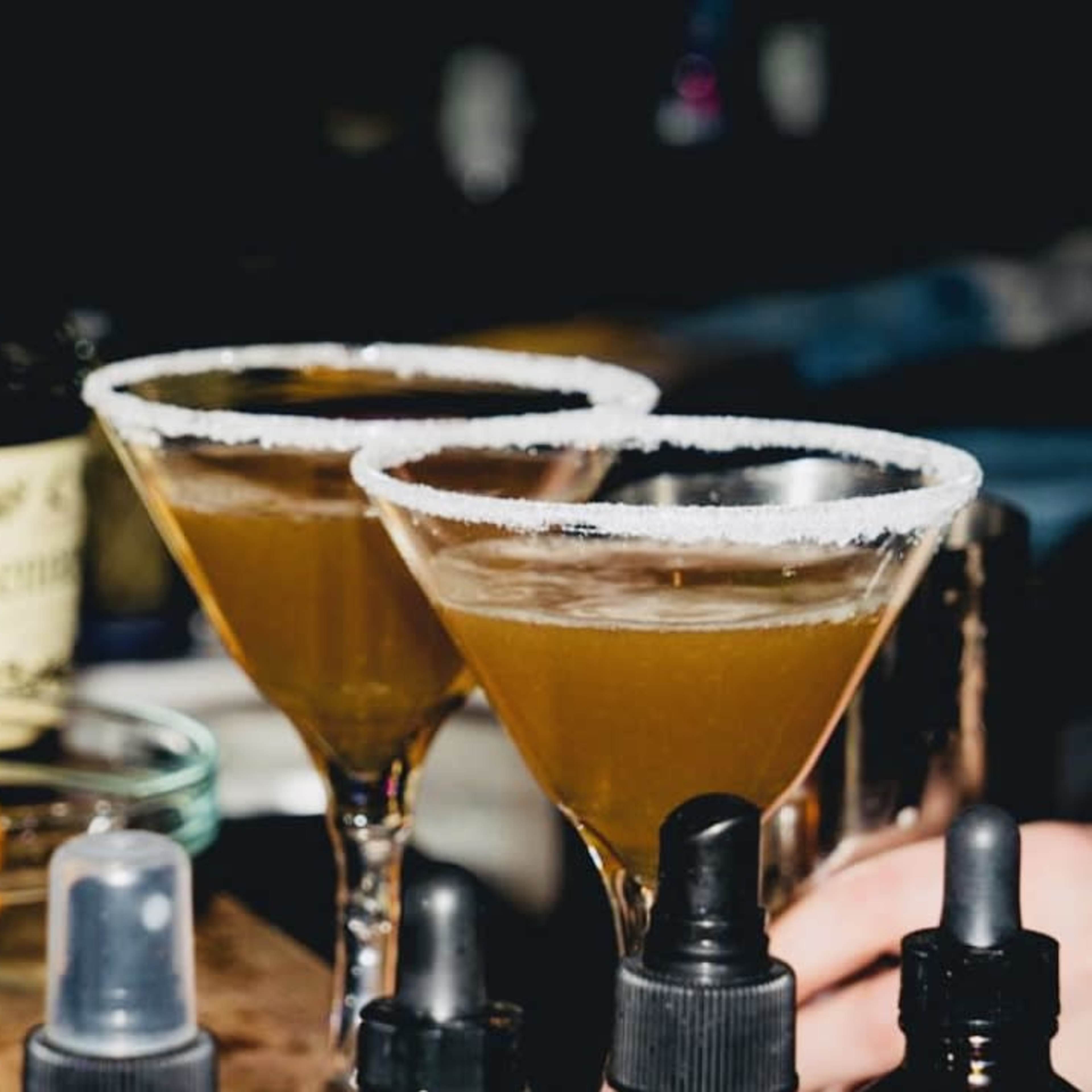 Two cocktails with salt rims are placed on a bar alongside a bottle of liquor and a orange slice.