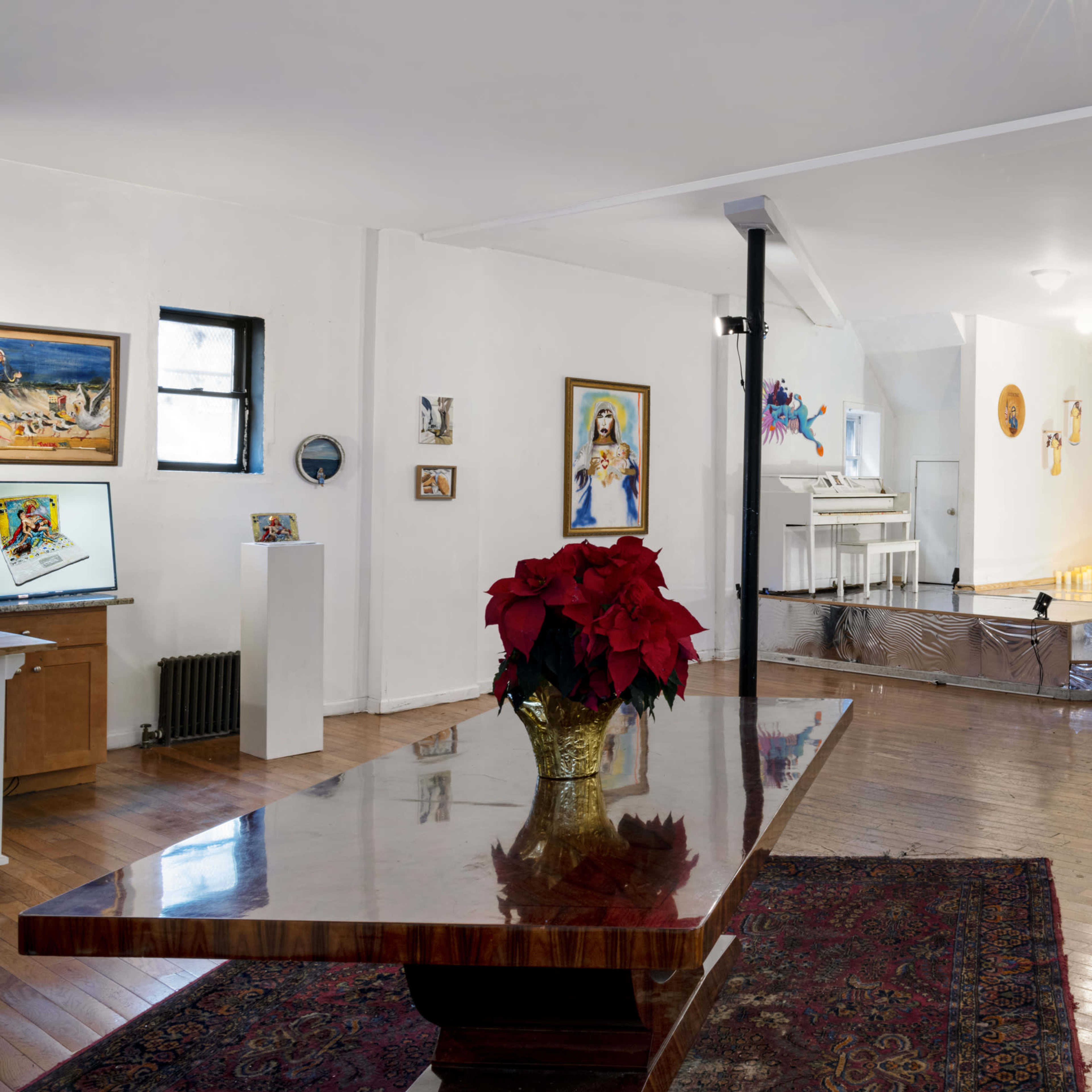 A spacious art gallery featuring a hardwood floor, various artworks on the walls, and a large table with a flower arrangement at the center.