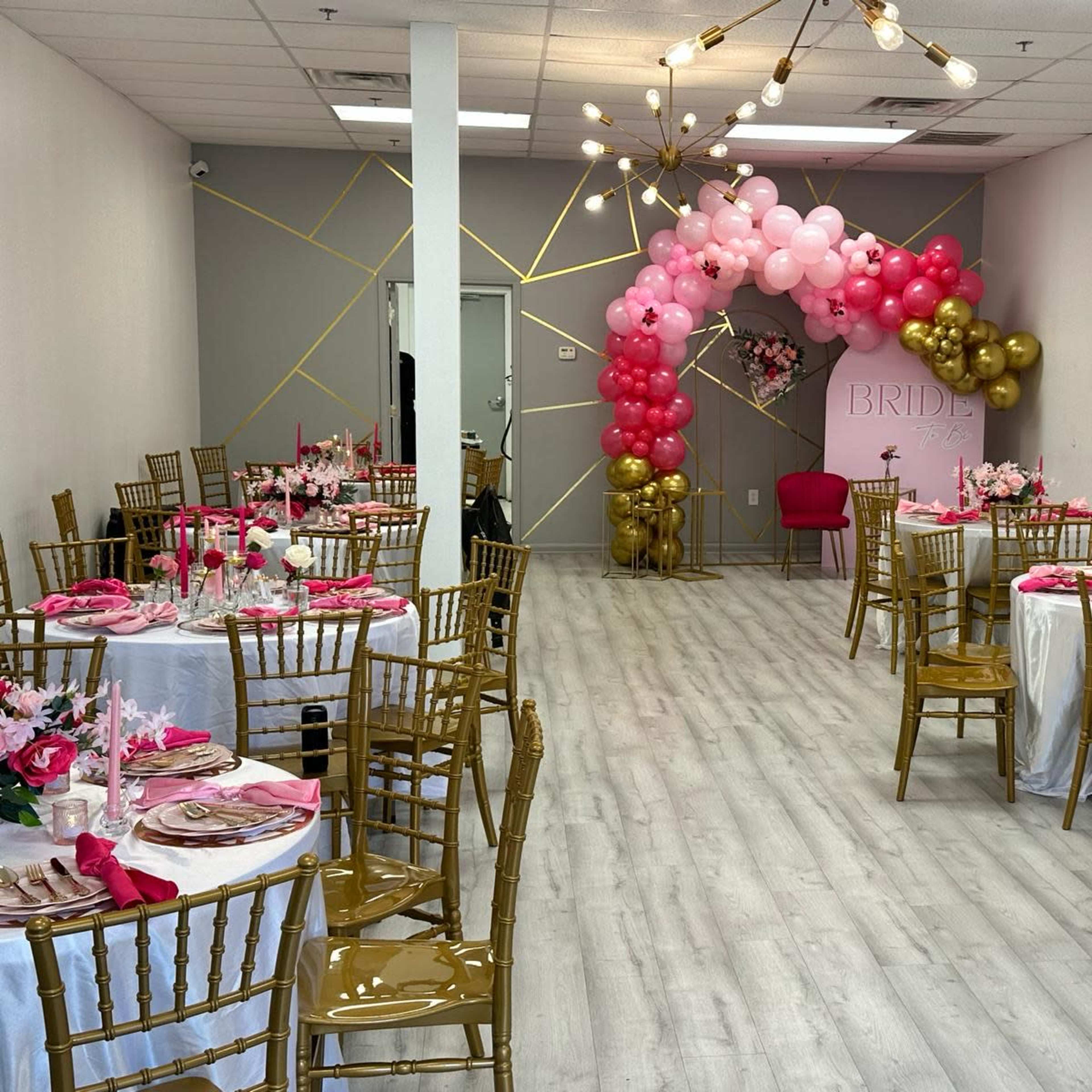 A decorated event space with round tables set for a celebration, featuring gold chairs, pink and white table settings, and a balloon arch.