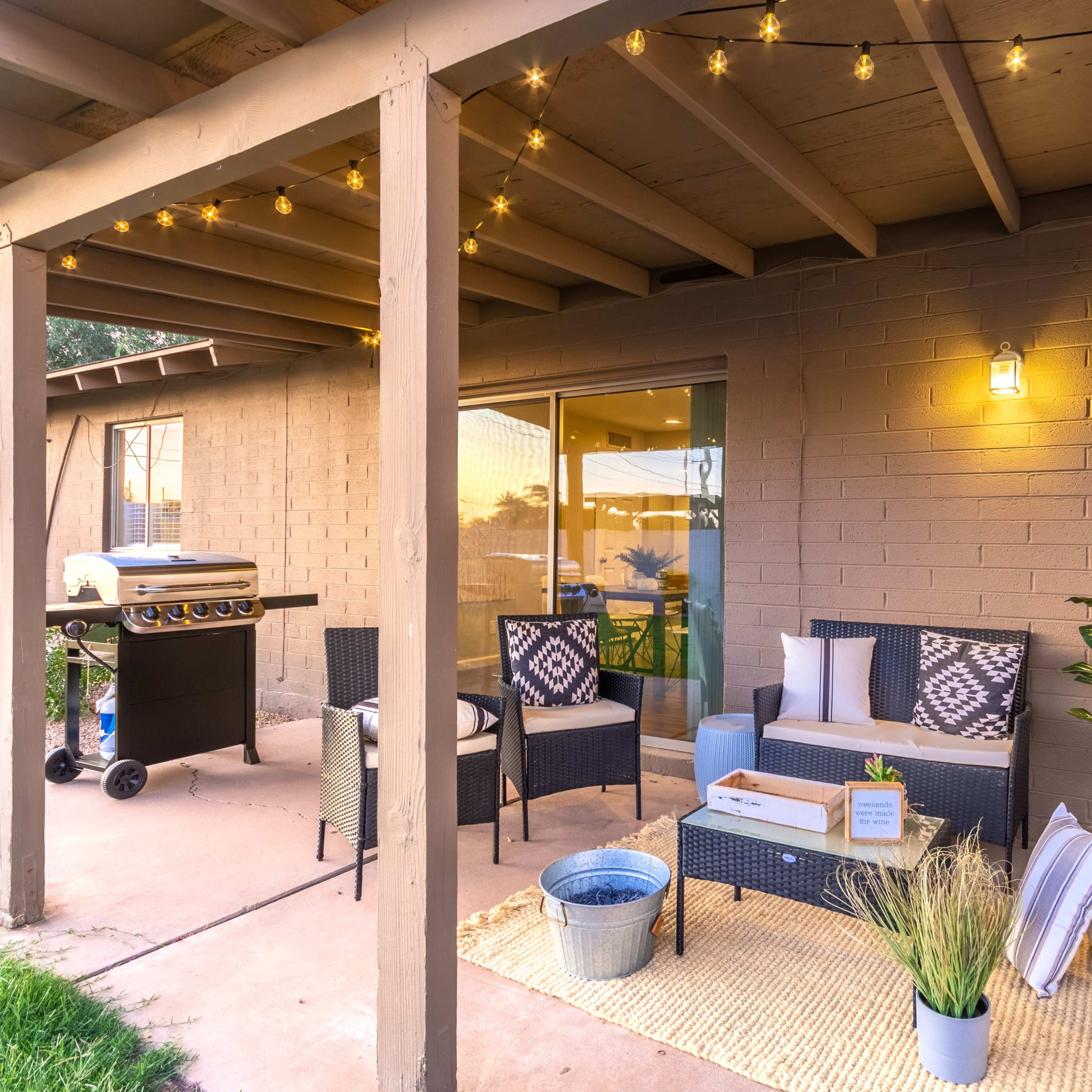 A patio area featuring a seating arrangement with cushions, a grill, string lights overhead, and decorative plants.