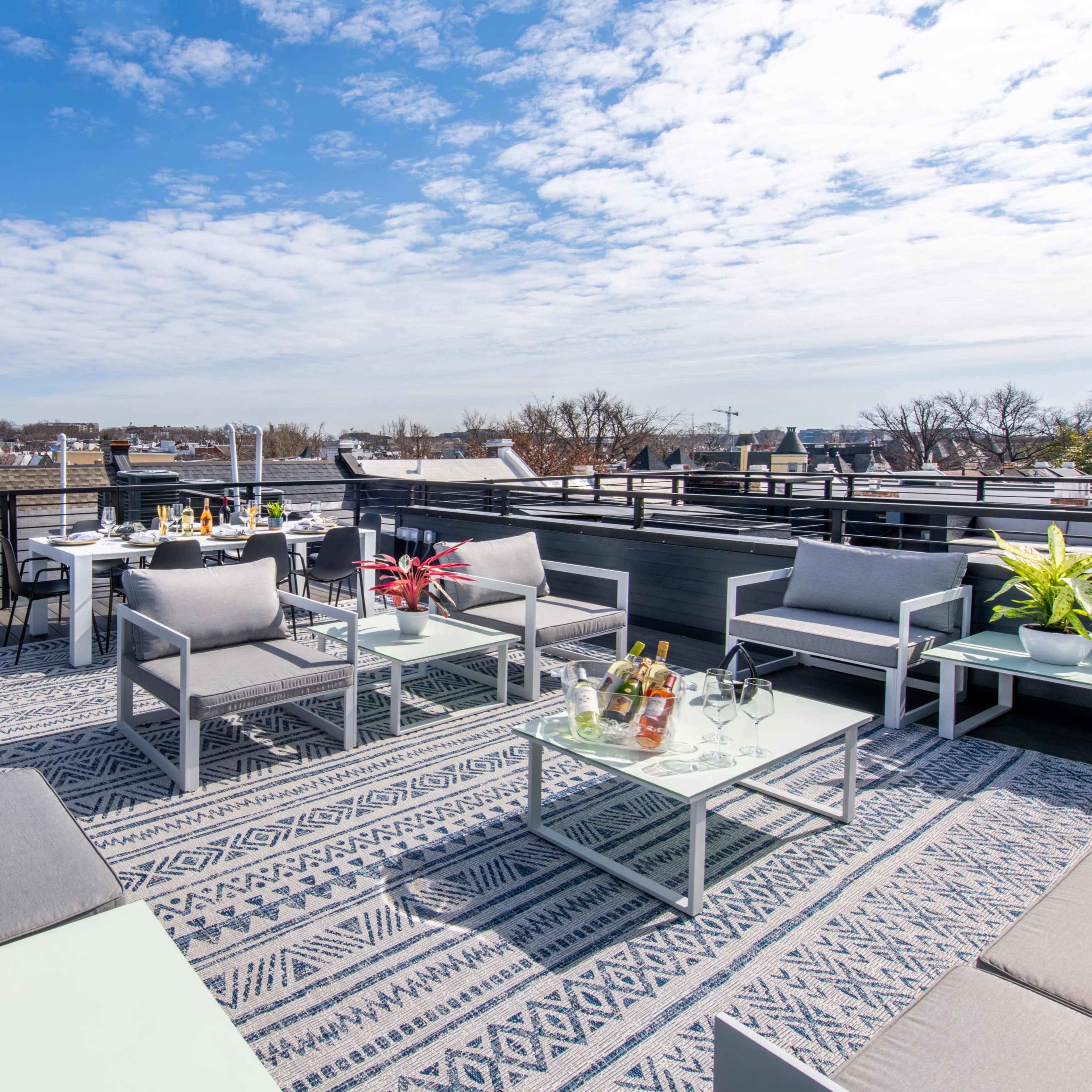 A rooftop terrace furnished with several seating areas, a decorative rug, and an assortment of beverages on tables under a partly cloudy sky.