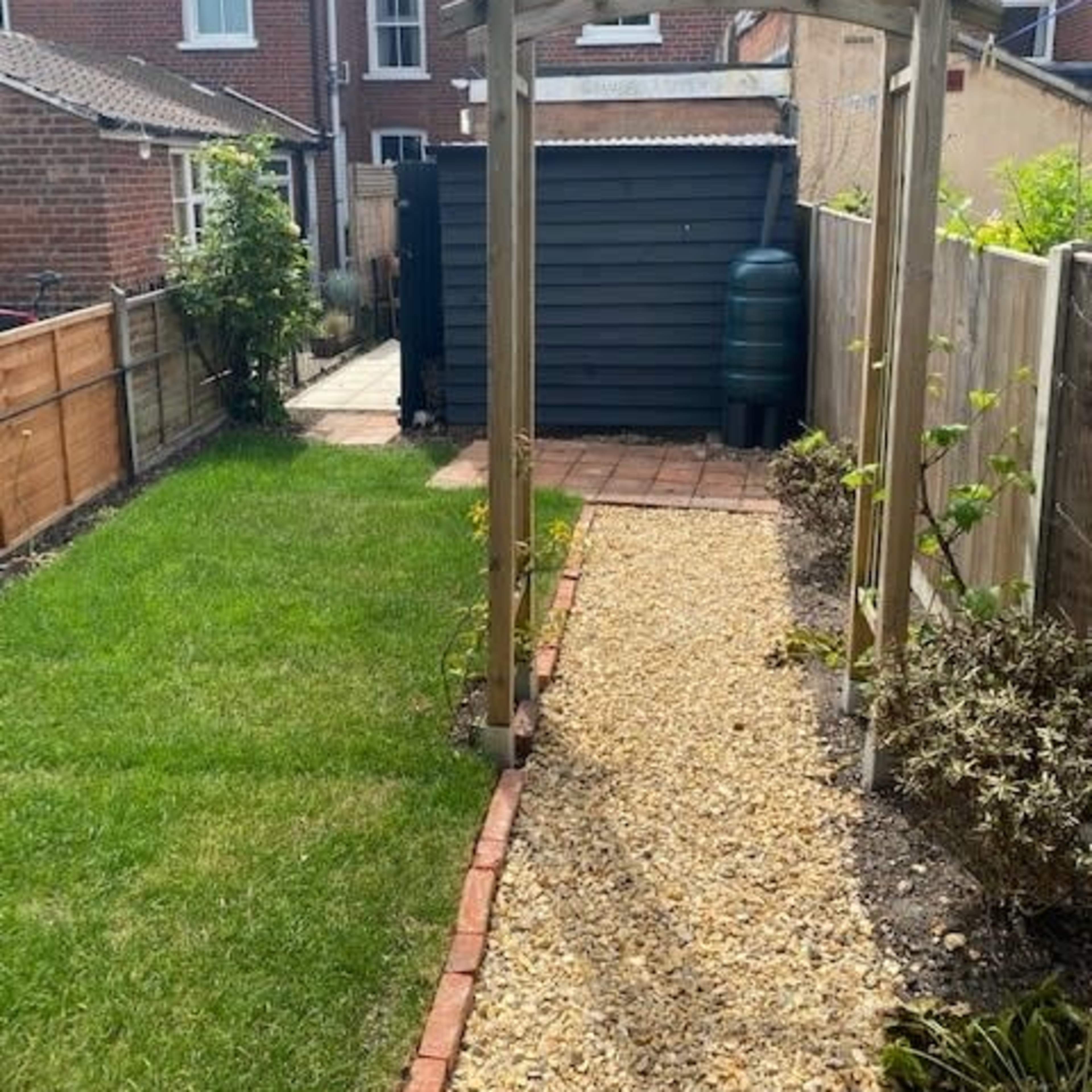 A garden path made of gravel leads to an archway with a wooden frame, surrounded by grass and shrubs, with a shed and water barrel in the background.