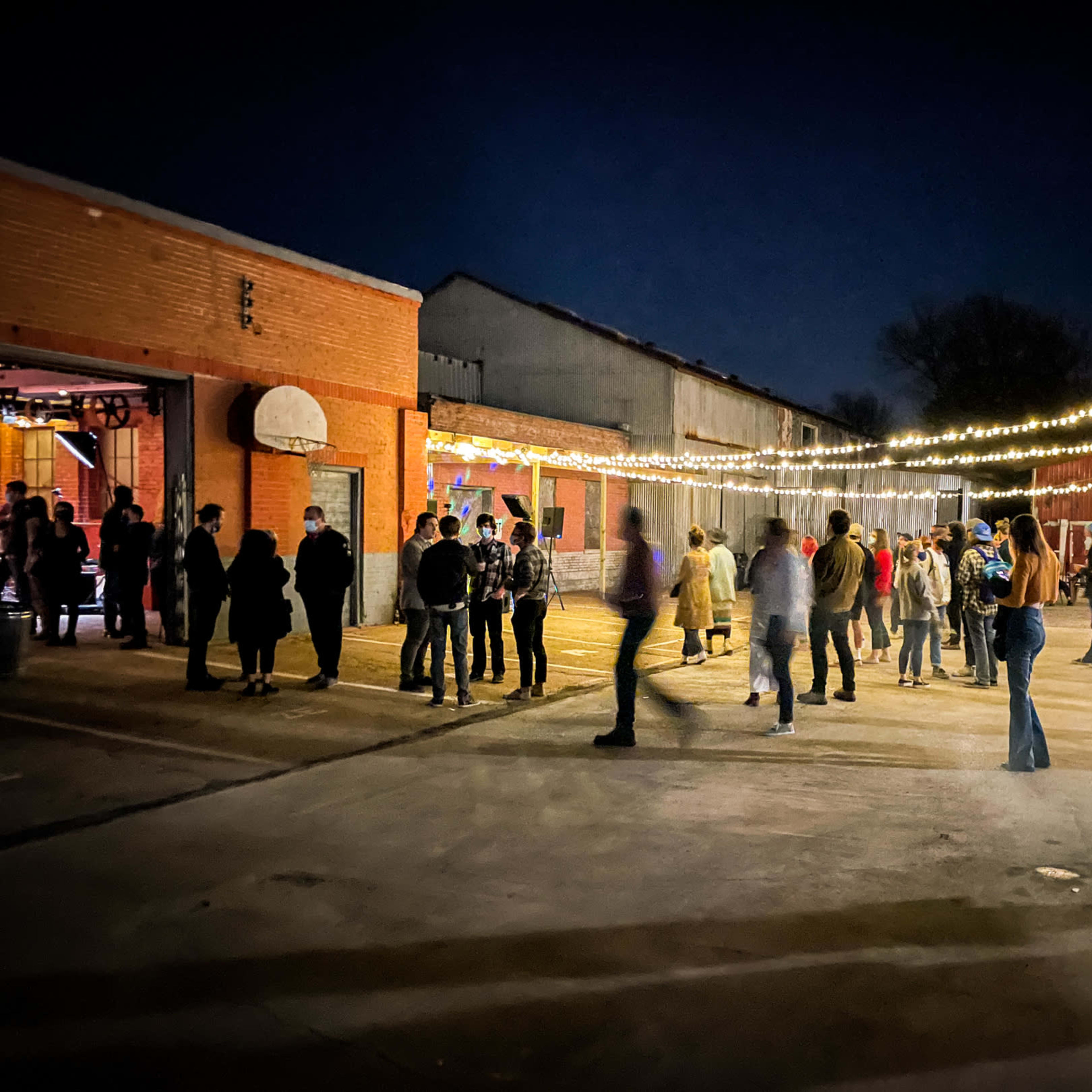 A crowd of people stands in a lit outdoor area with string lights above, surrounded by brick buildings at night.