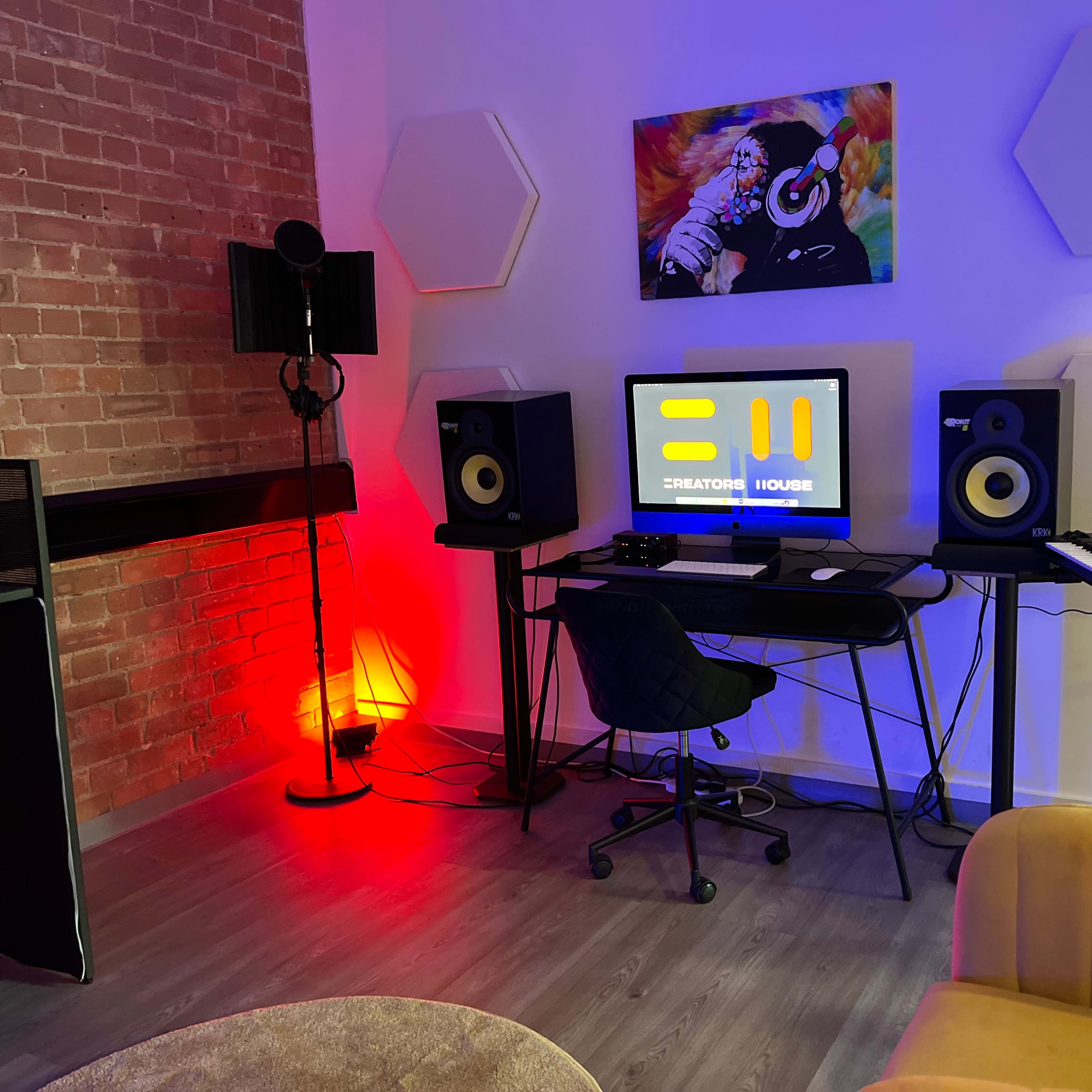 A modern room set up as a music recording studio, featuring a desk with a computer, speakers, a keyboard, and colorful lighting against a backdrop of exposed brick.