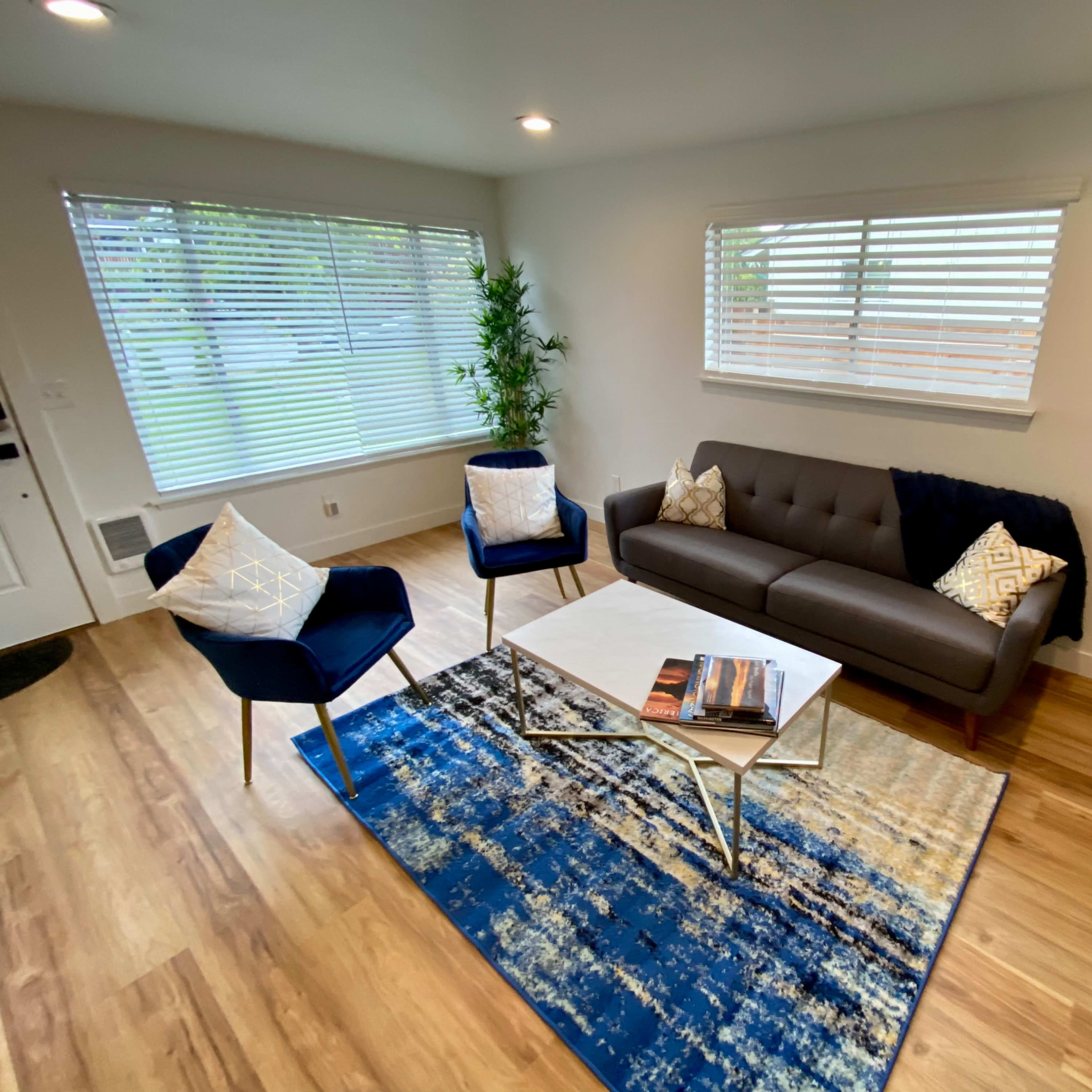 A modern living room featuring a gray sofa, two dark blue chairs, a coffee table with a magazine, and a large rug, all illuminated by natural light from the windows.