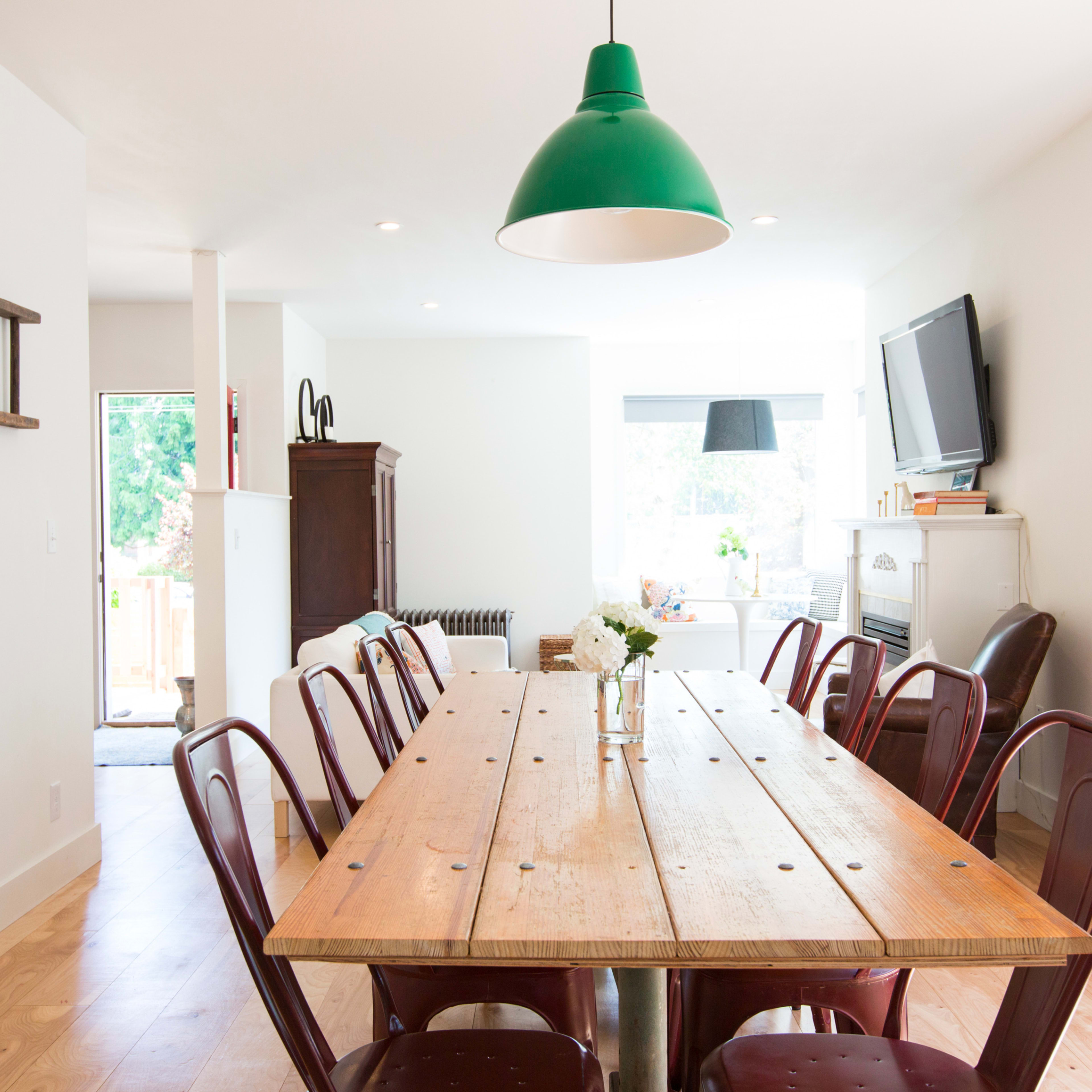 A long wooden dining table with metal chairs is set in a bright, minimalistic room featuring a green pendant light and a large window.